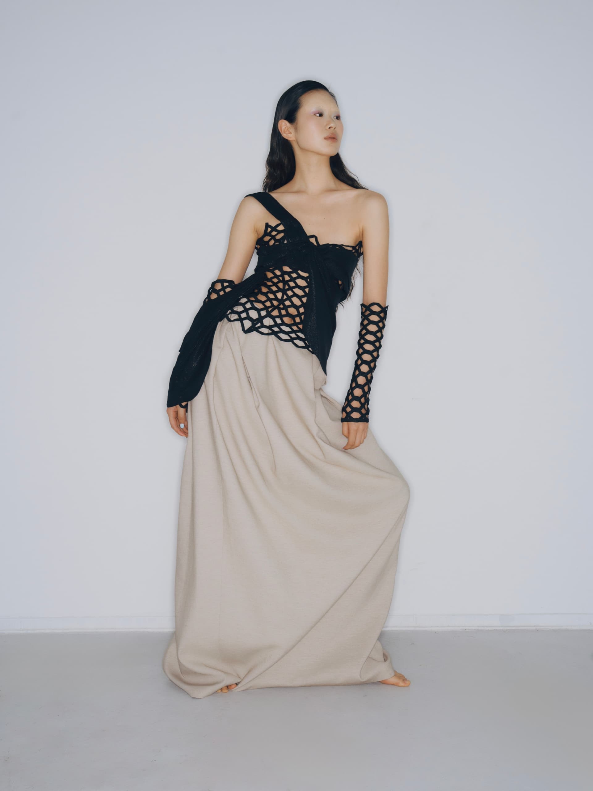 Fashion model in stylish black mesh top and beige maxi skirt against white backdrop.