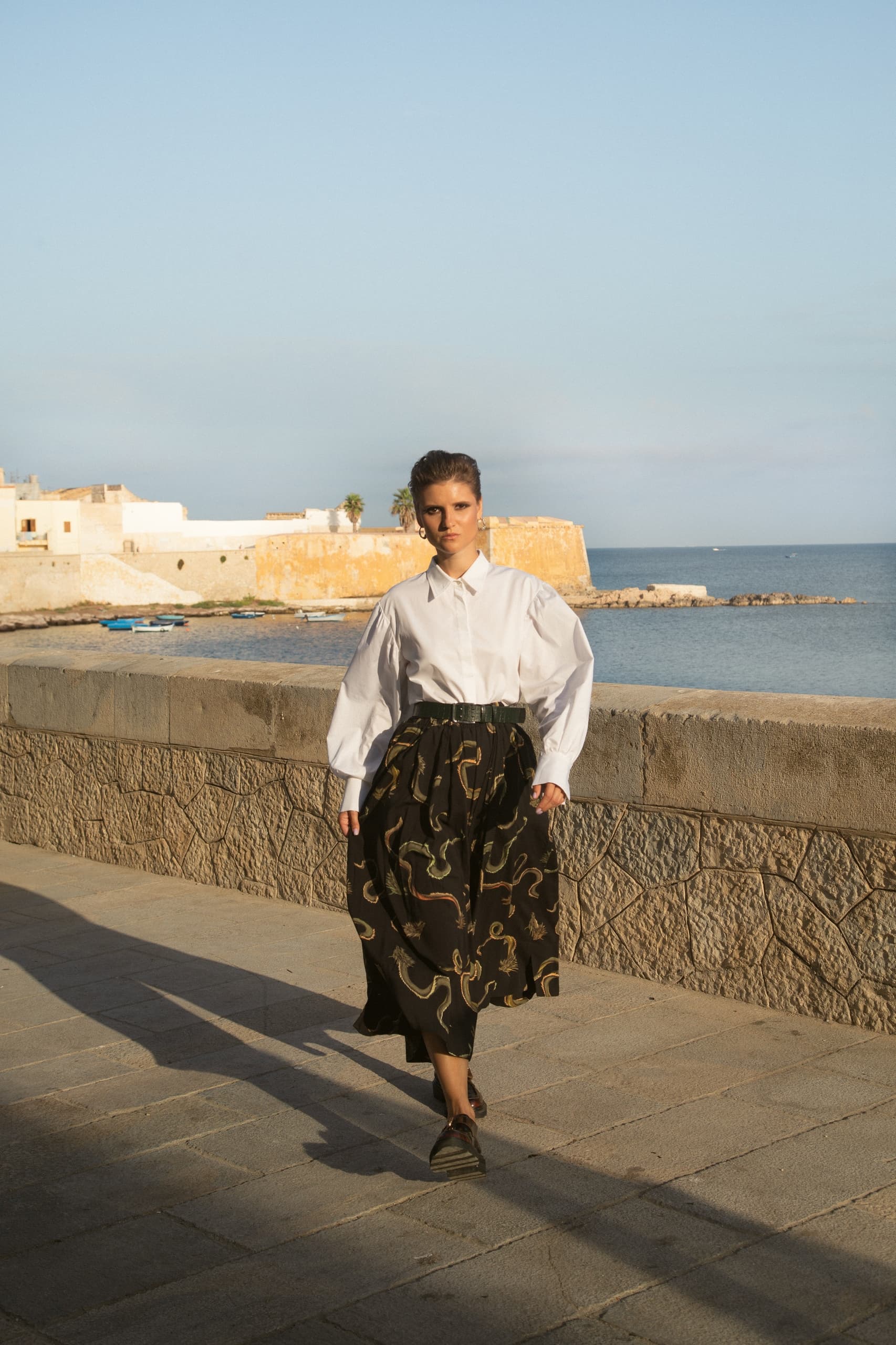 Woman in white blouse and patterned skirt walking seaside at sunset.