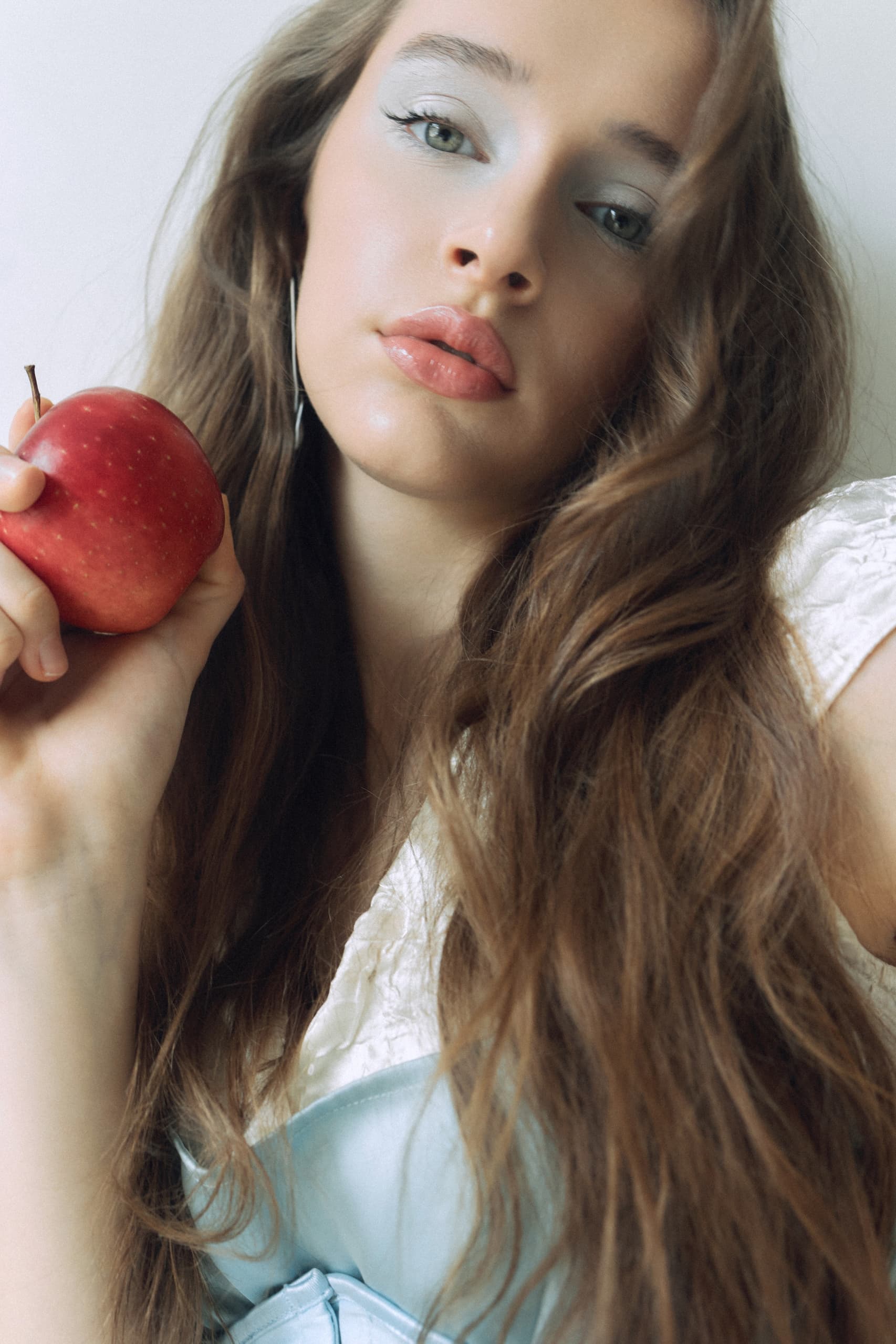 Woman with long hair holding a red apple, wearing pastel makeup and white top.