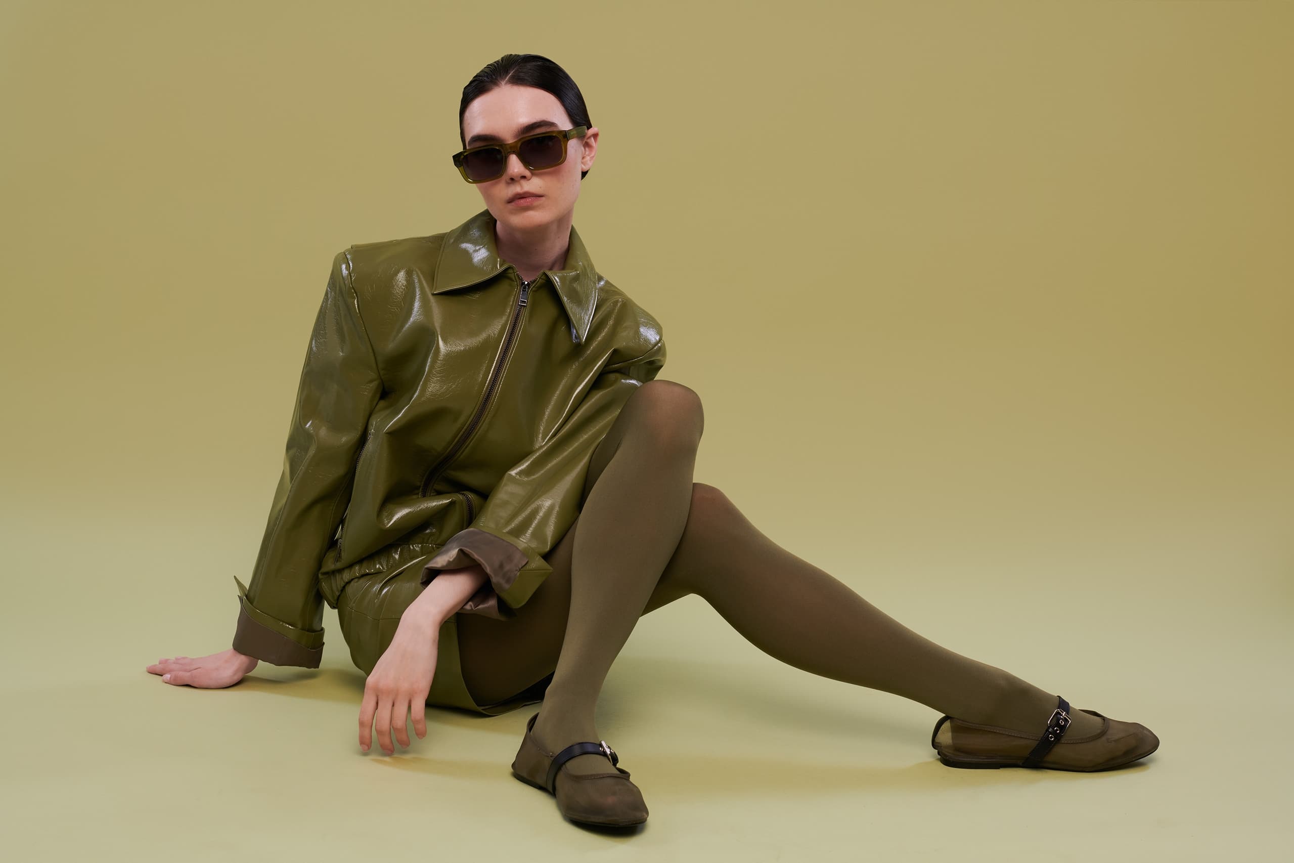 Fashionable model in green leather coat with sunglasses poses on a neutral backdrop.