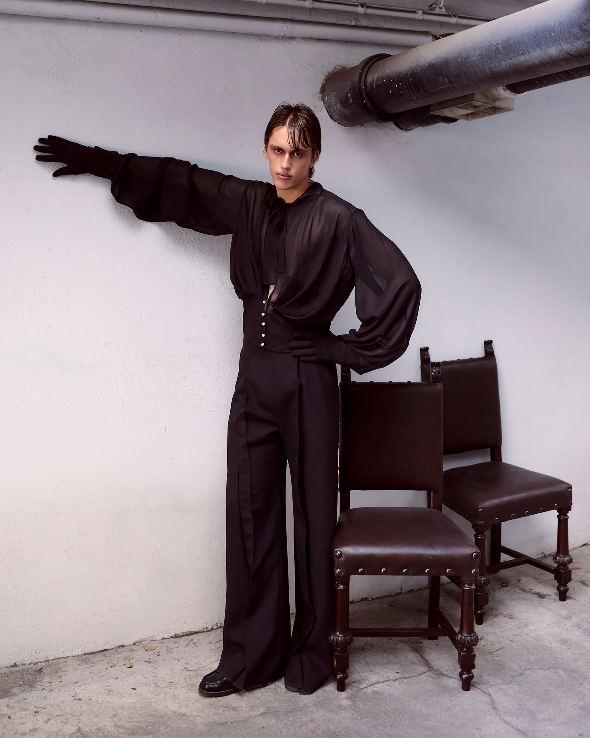 Person in dark, stylish outfit poses by chairs in an industrial-themed room.