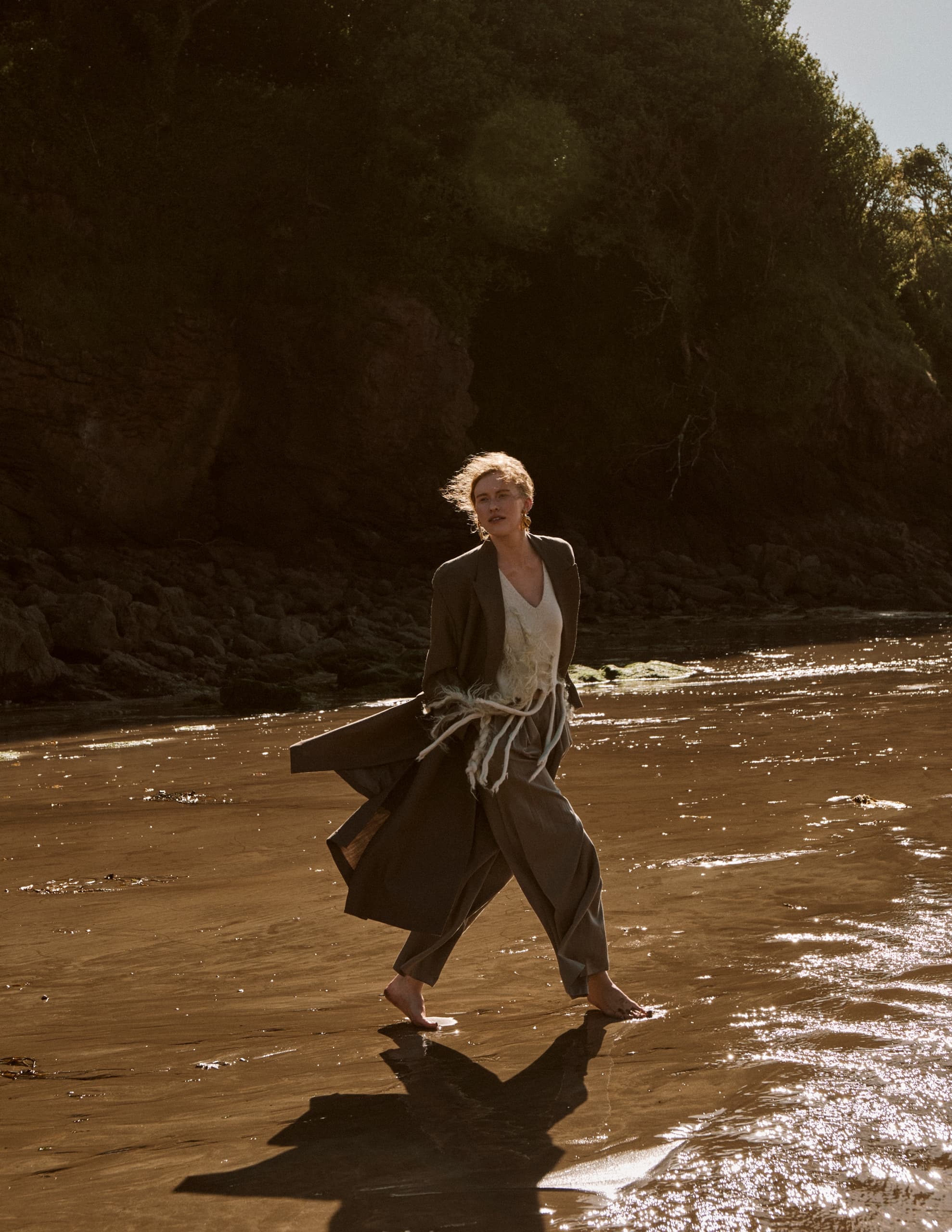 Woman in flowing outfit walking on sunlit beach with rocky backdrop.