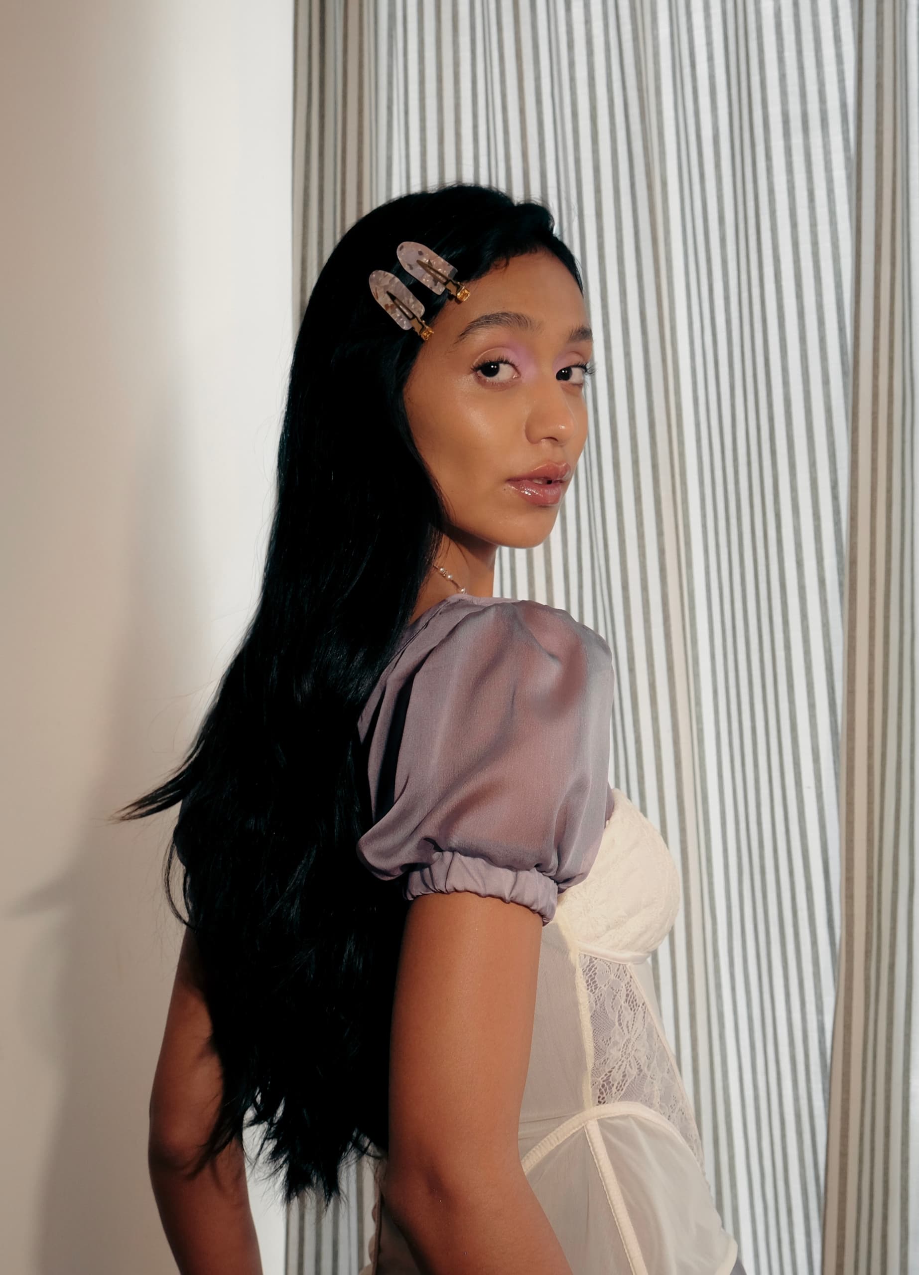 Woman in sheer purple top with hair clips, standing by striped curtains.