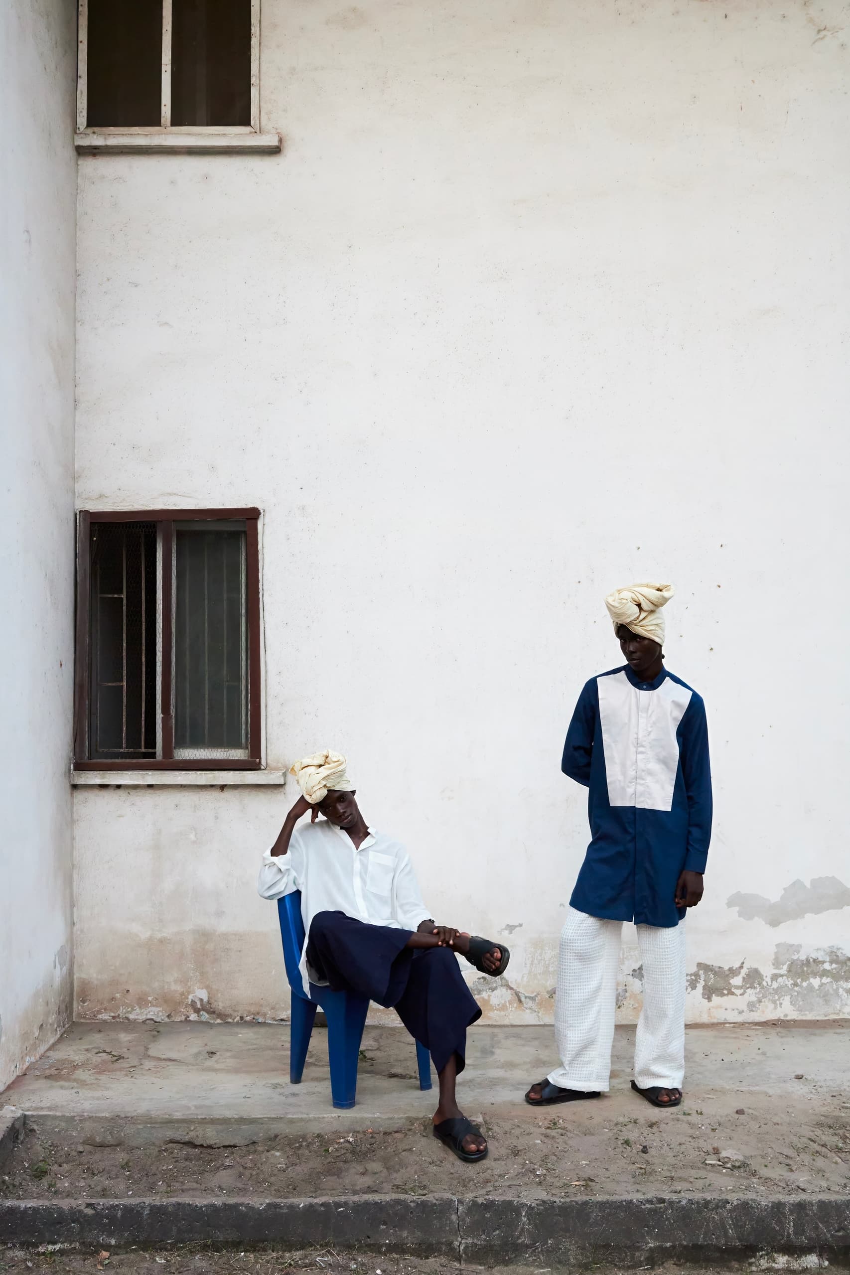 Two men in stylish attire with turbans outside, one seated, one standing. Minimalist scene.