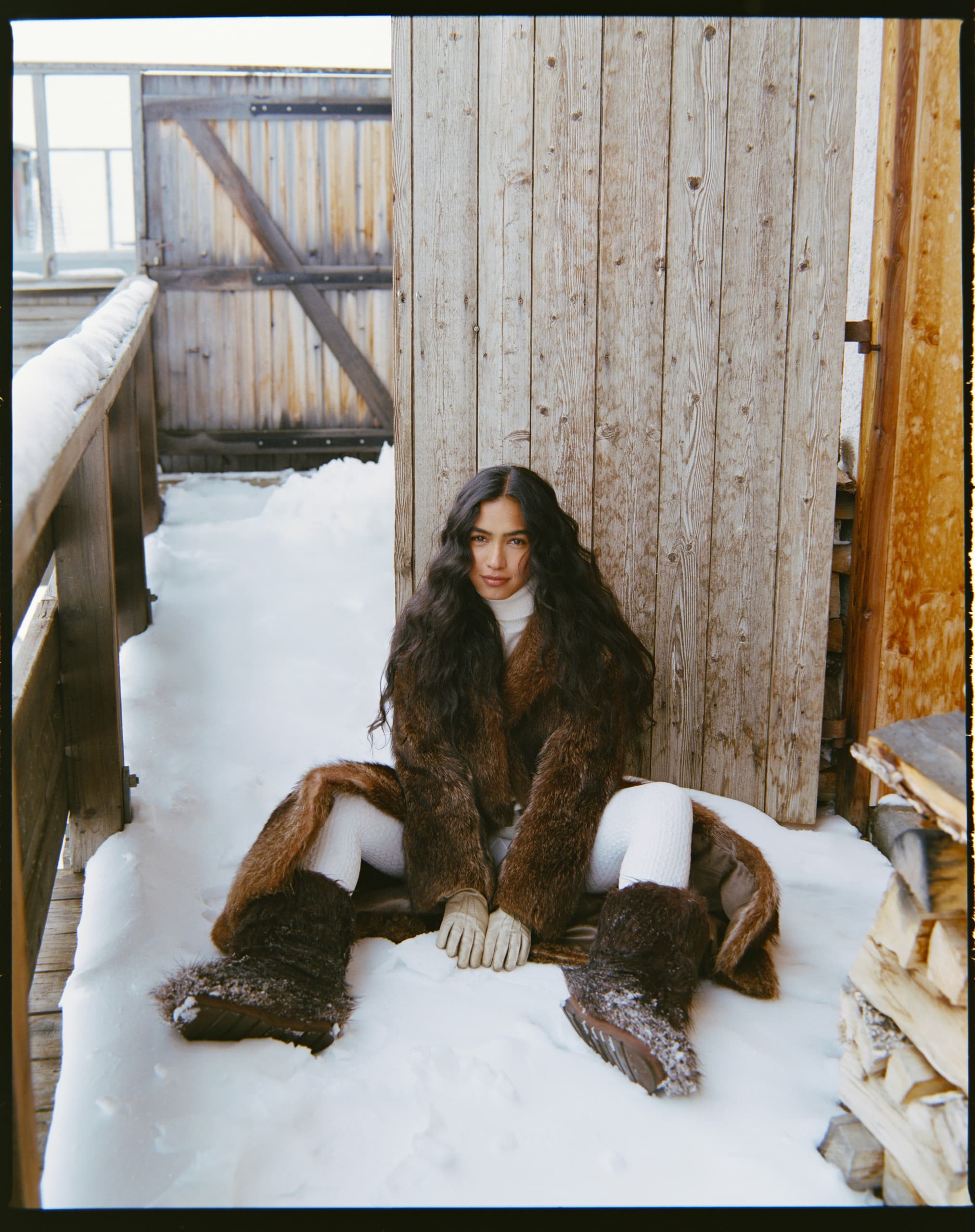 Woman in fur coat sitting in snowy cabin setting.