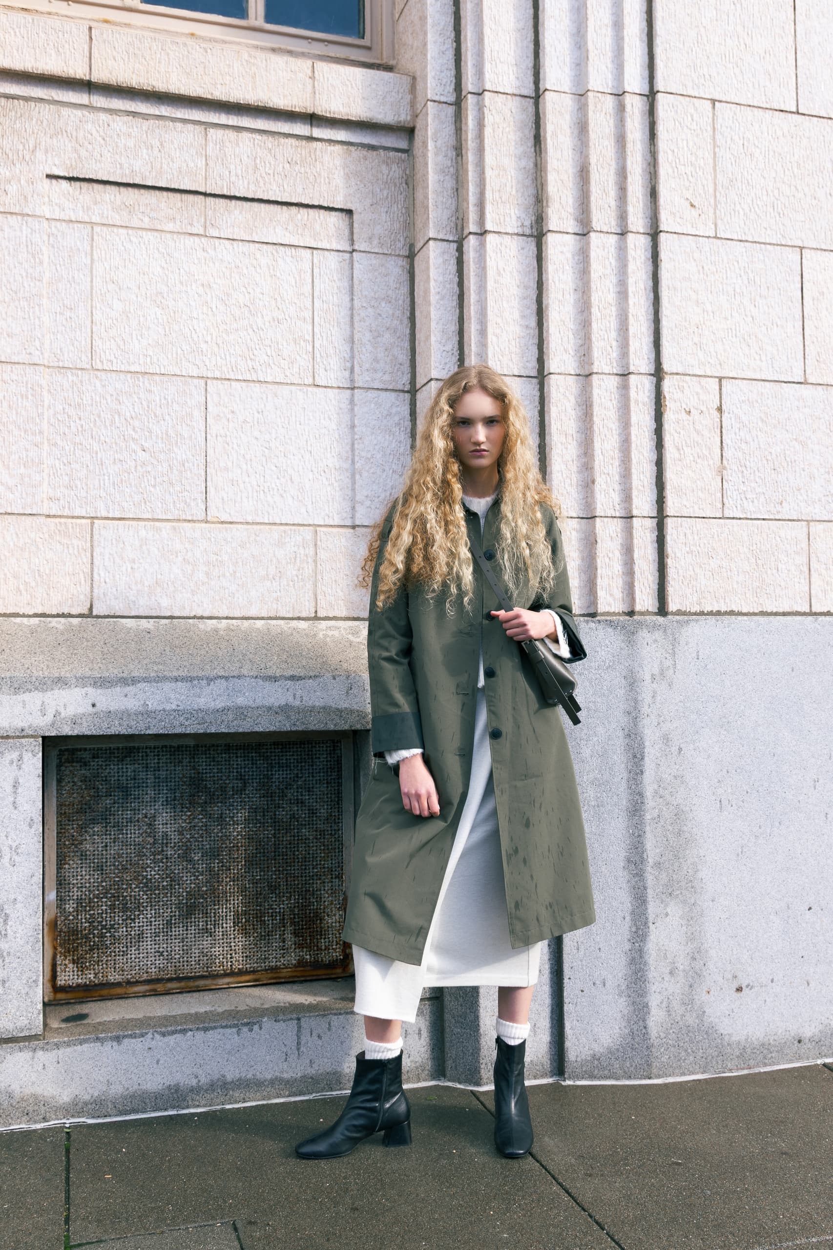 Woman in a green coat standing against a textured stone wall.