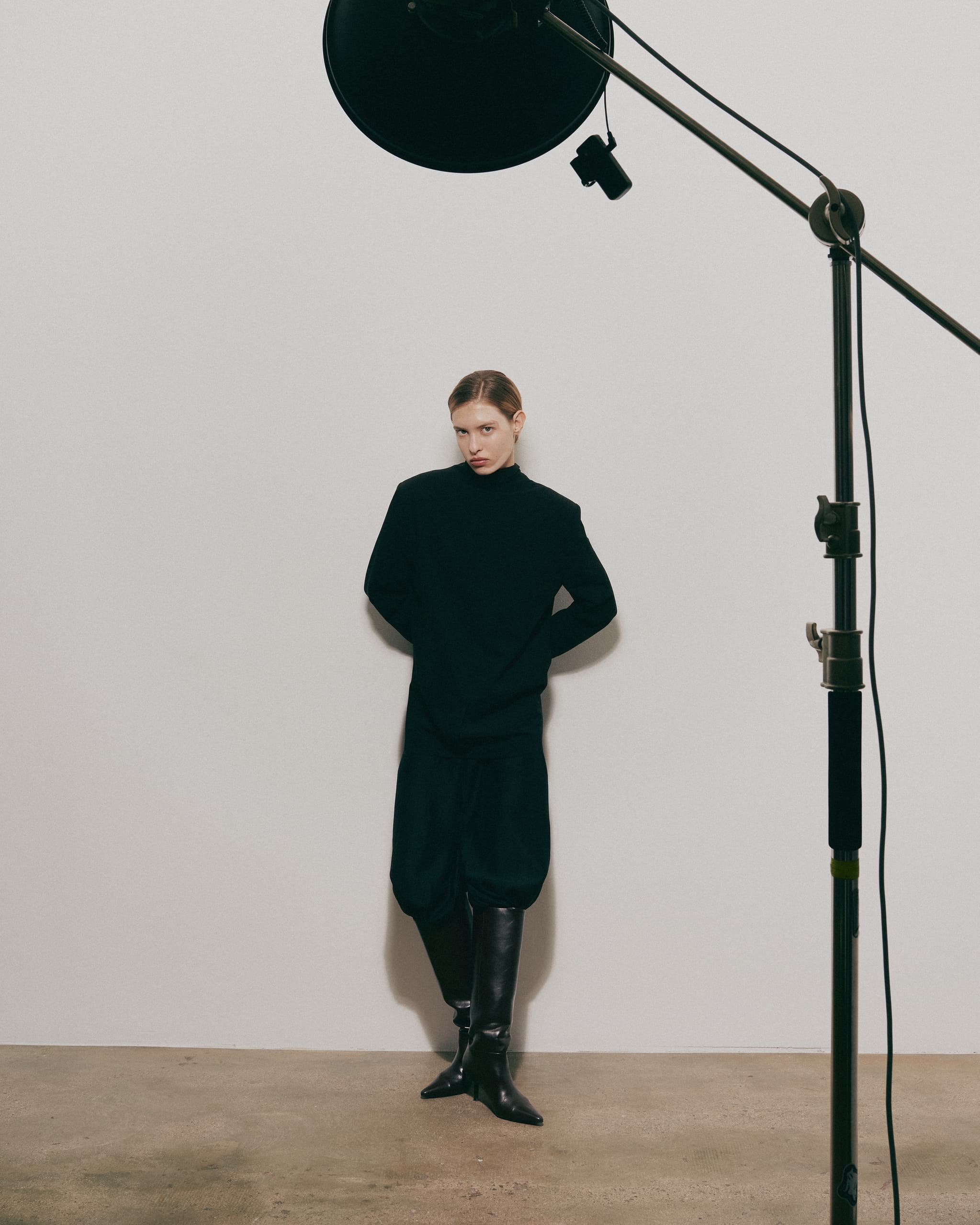 Model in black outfit poses in a studio under soft lighting.