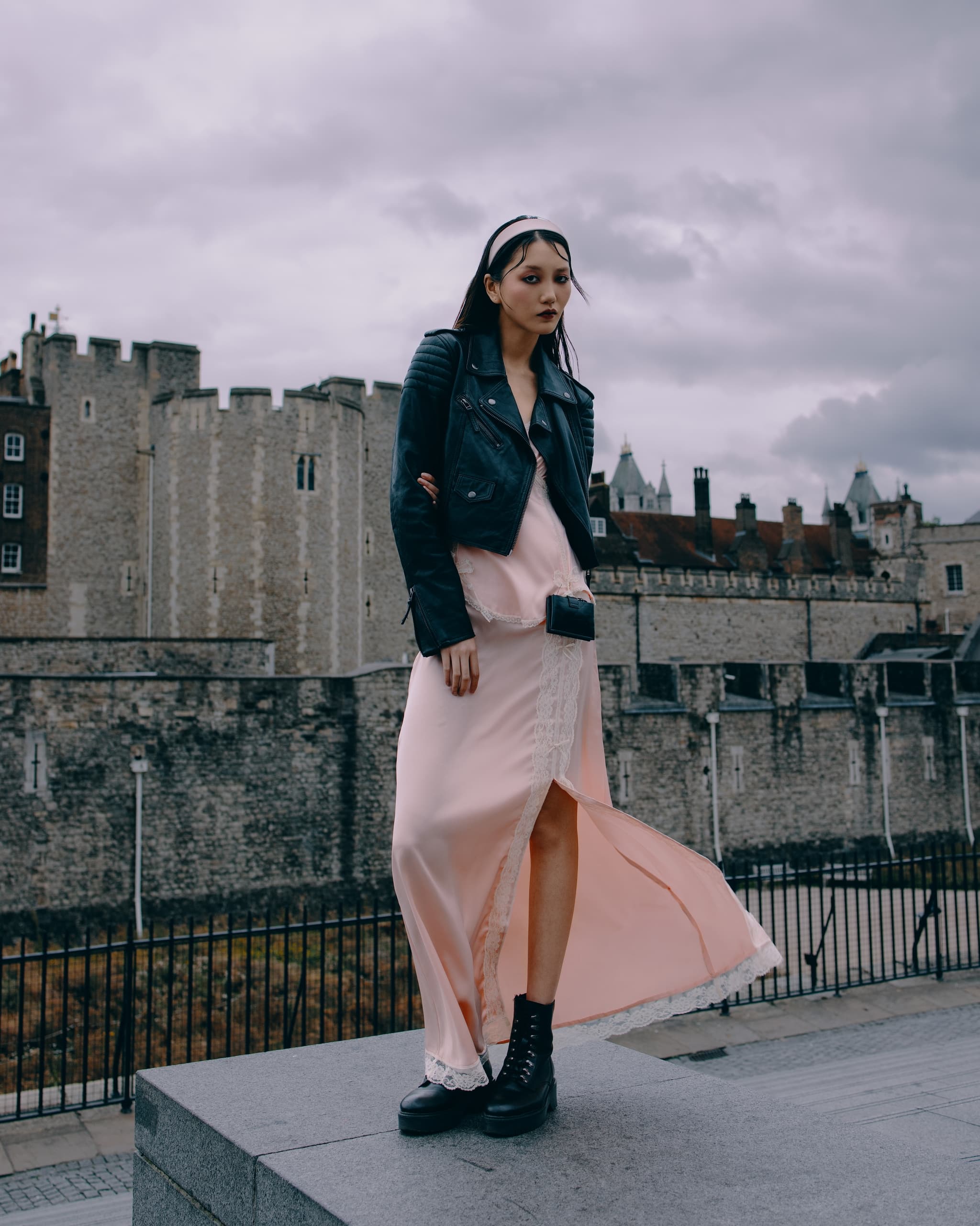 Fashionable woman in pink dress and leather jacket by a historic stone building.