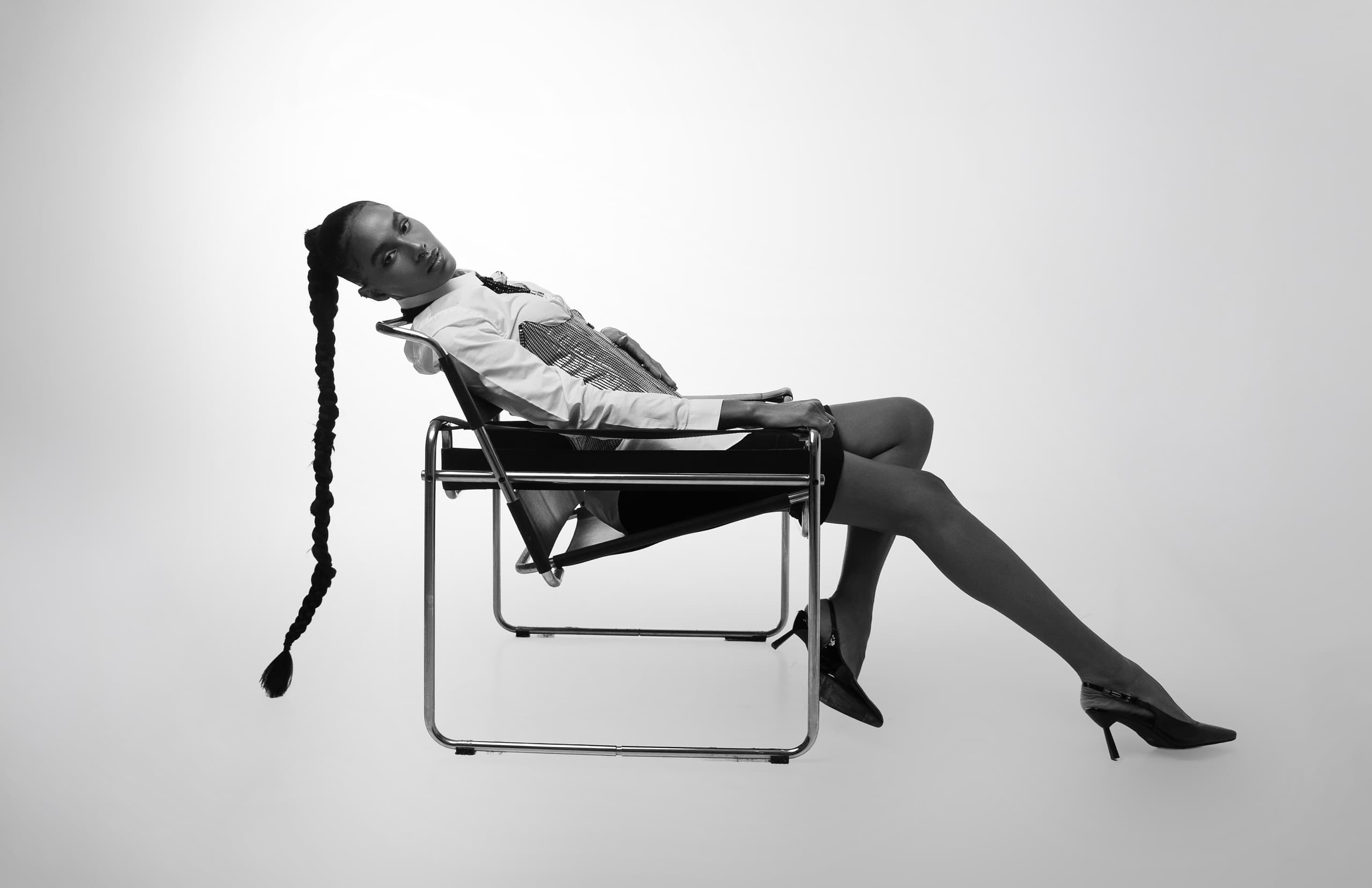 Person in stylish pose on a chair, showcasing long braid and modern fashion.