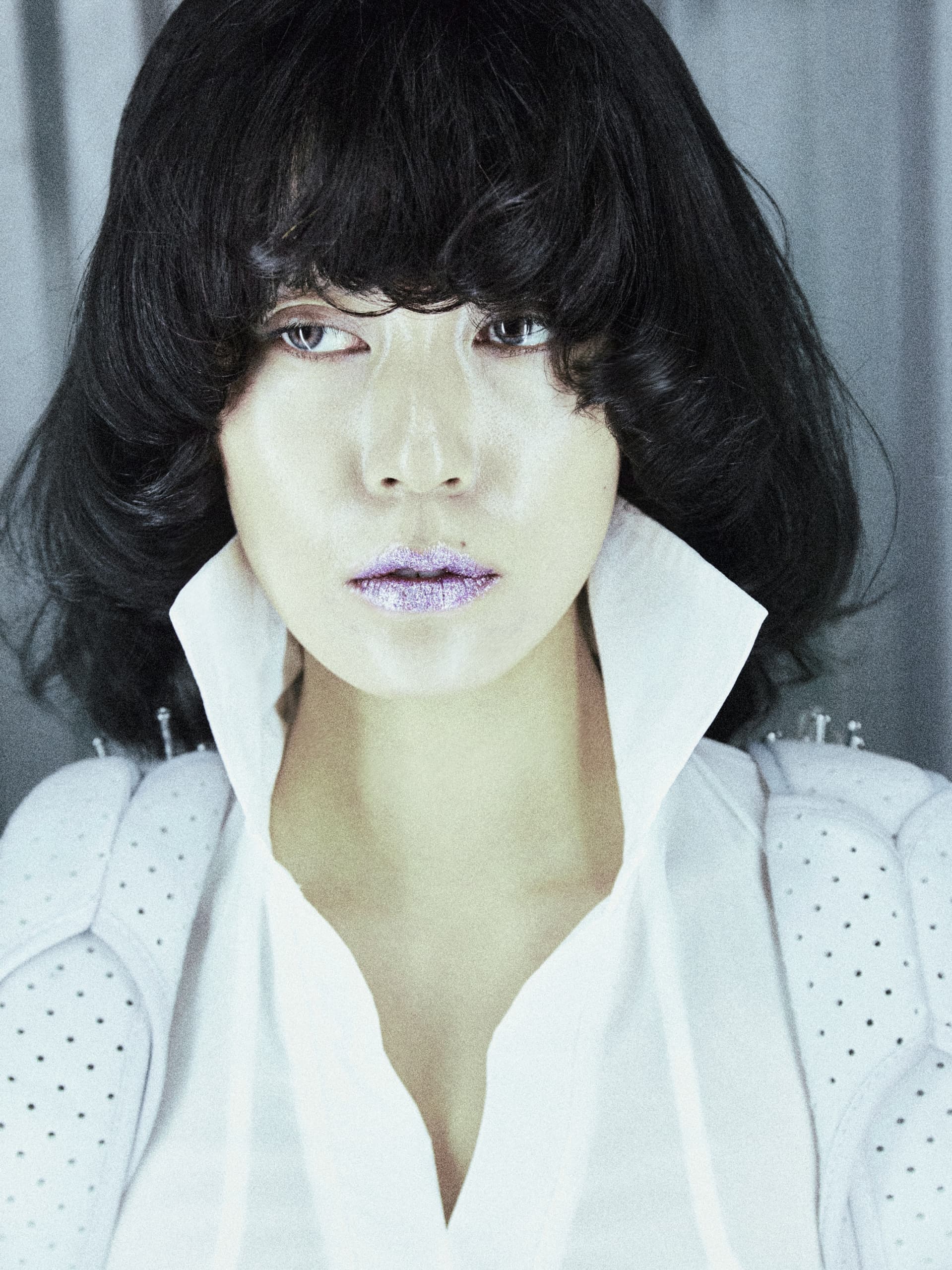 Fashion model with bold hairstyle and shimmering lips in a high-collared white outfit.