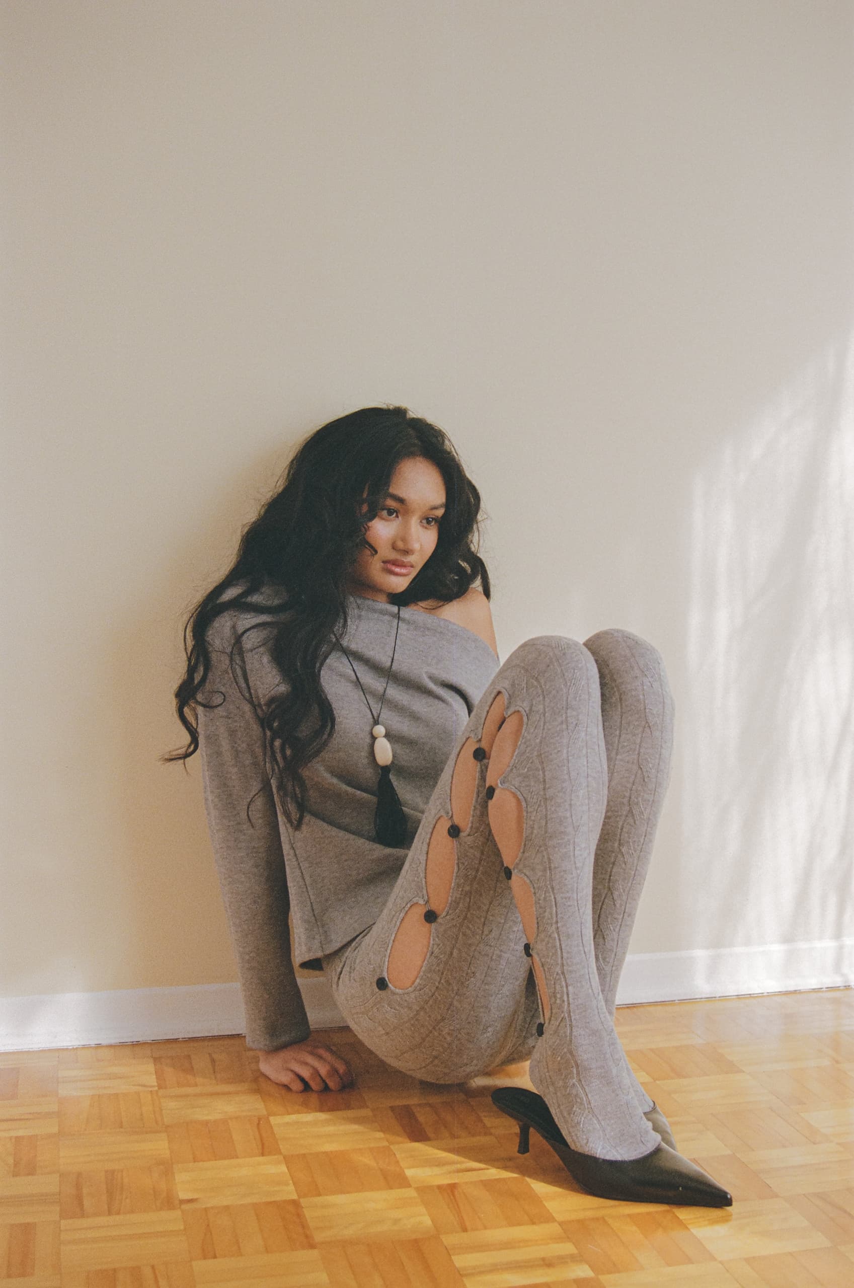 Woman in stylish gray outfit, seated on hardwood floor, with natural light.