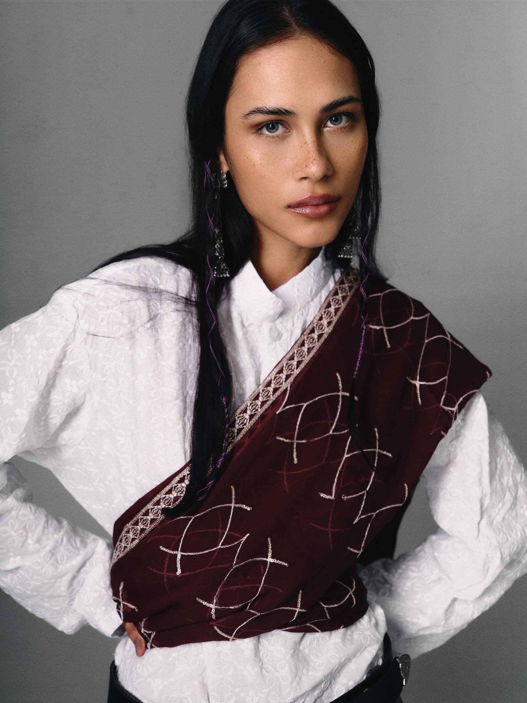Woman in embroidered scarf and white blouse, posing confidently with hands on hips.