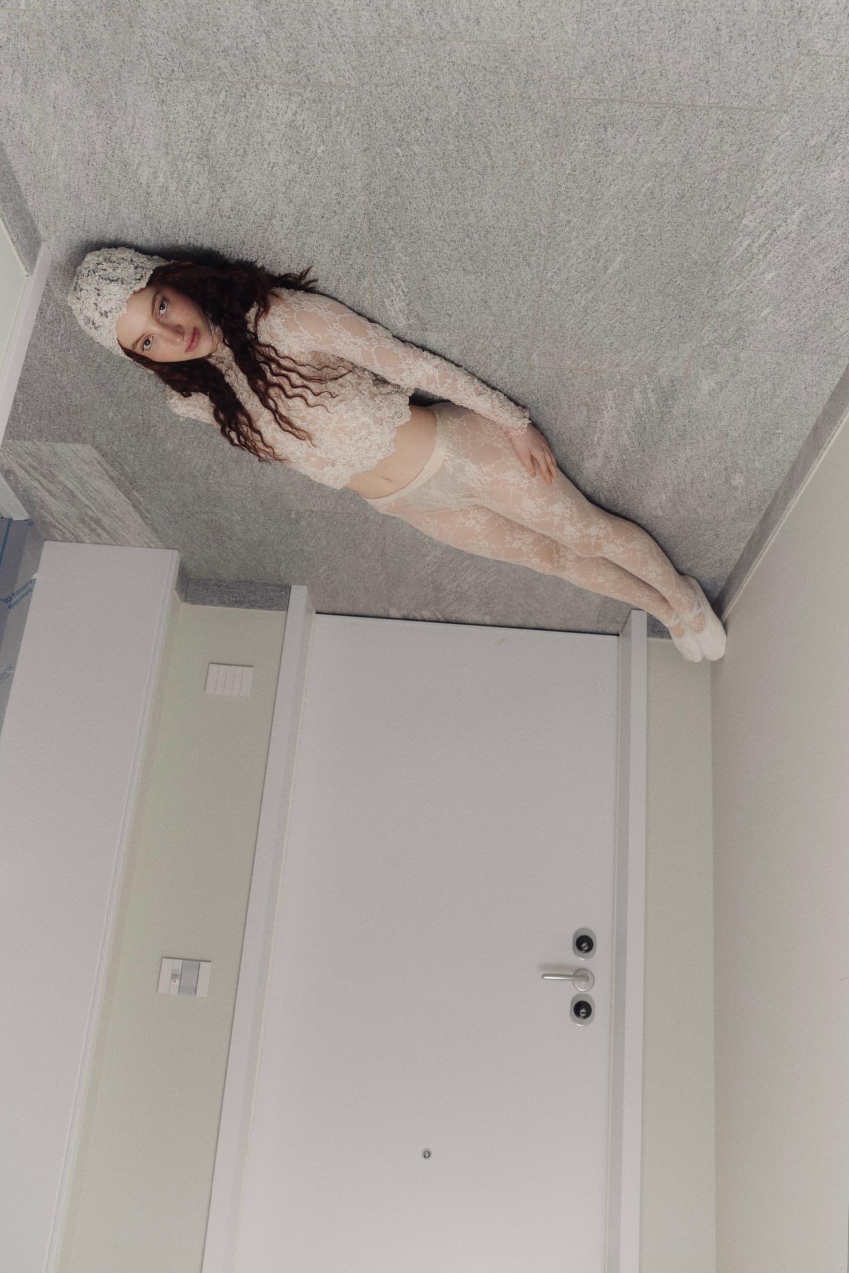 Person in lace outfit and knit hat lying on a floor near a white door.