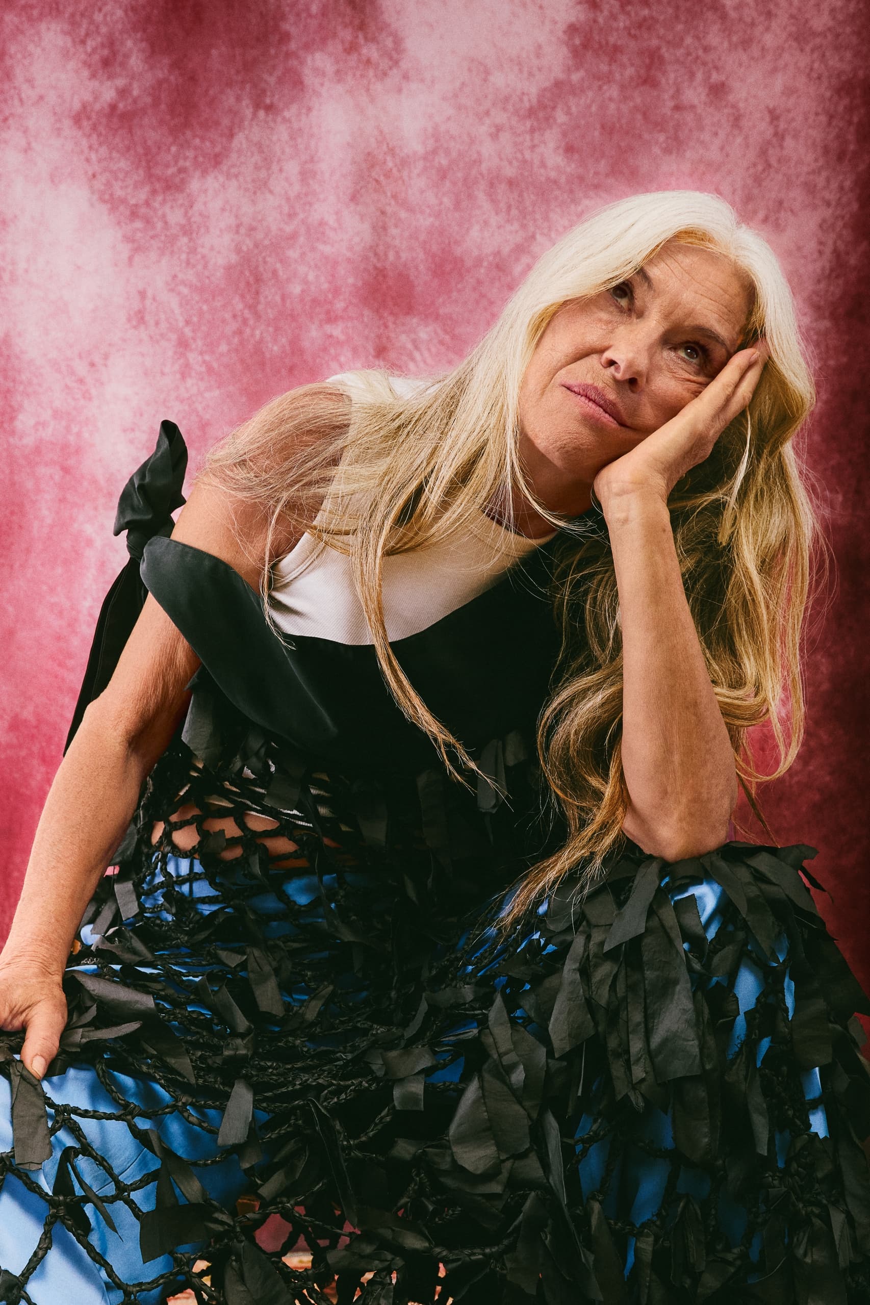 Woman with long hair and unique outfit, thoughtful pose against a pink background.