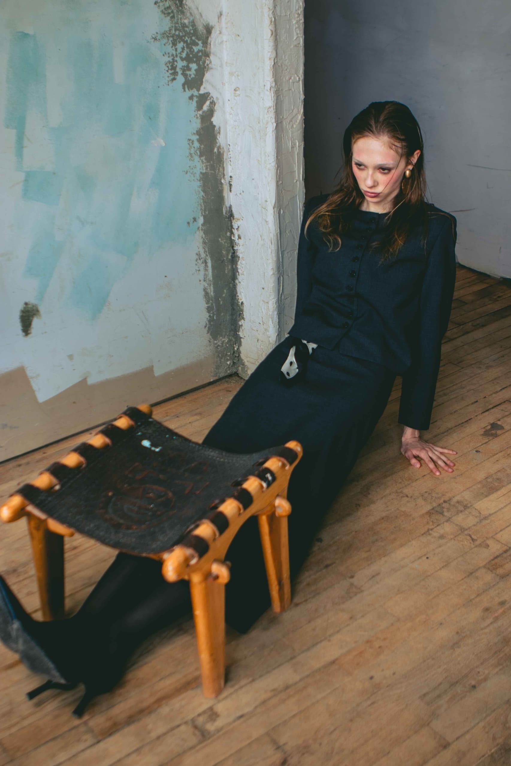 Woman in black outfit sits on wooden floor near rustic wall and small stool.
