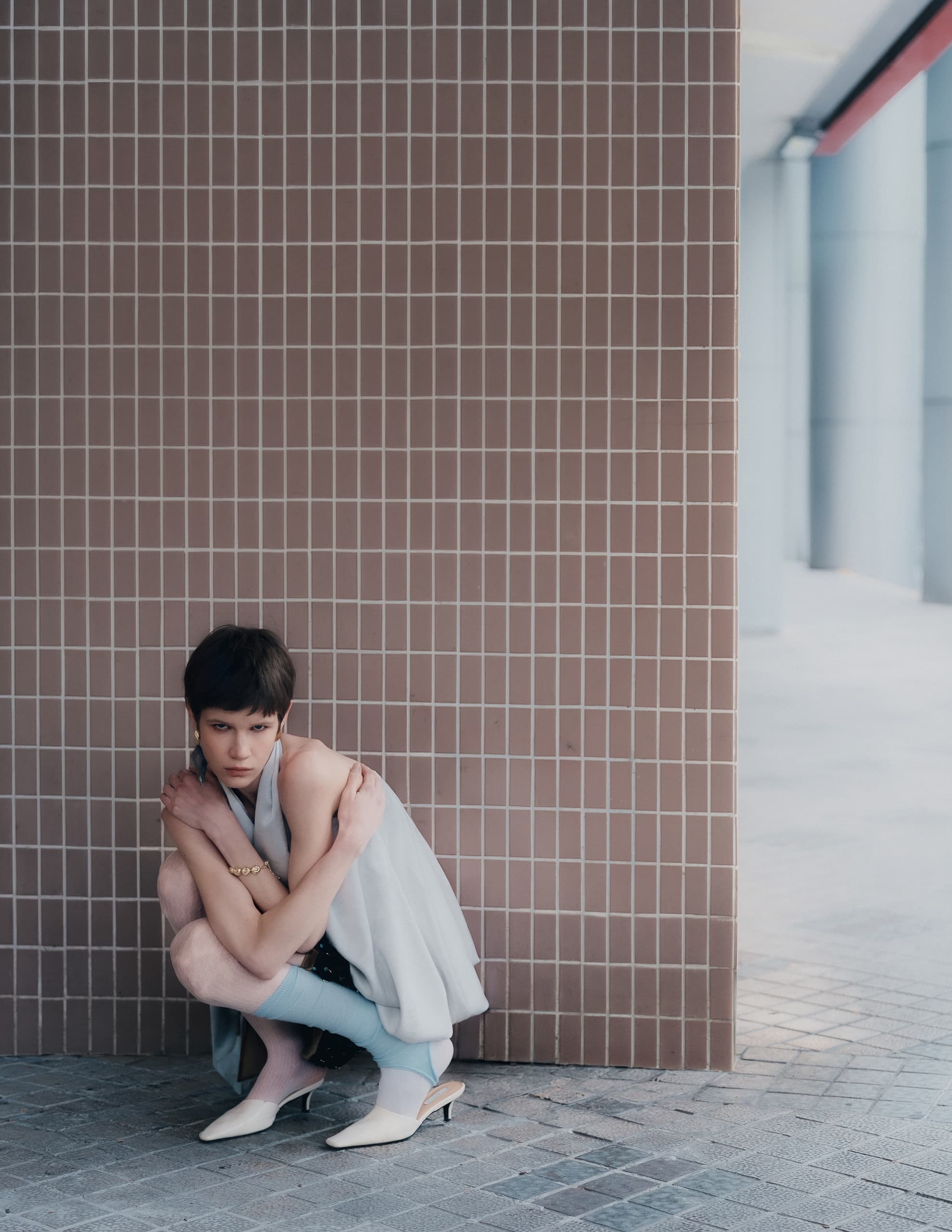 Fashion model crouching by tiled wall in stylish outfit and heels.