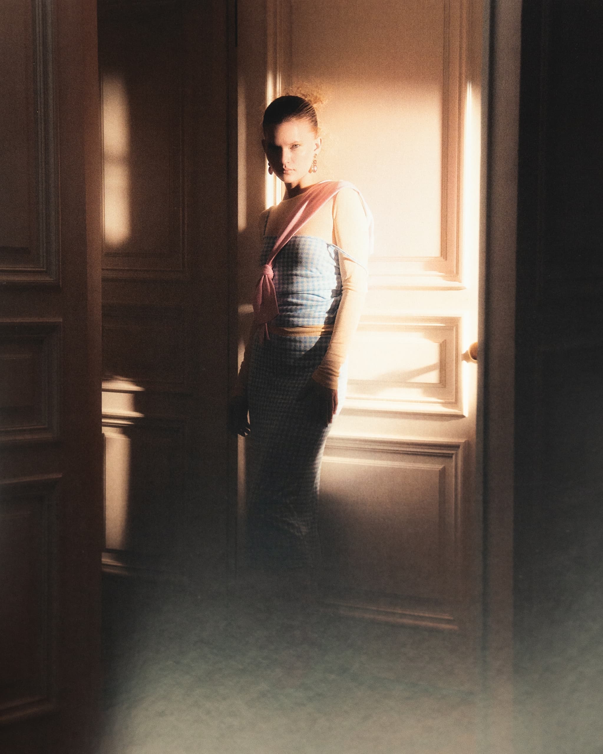 Woman in sunlight wearing stylish outfit, standing by a doorway in soft lighting.