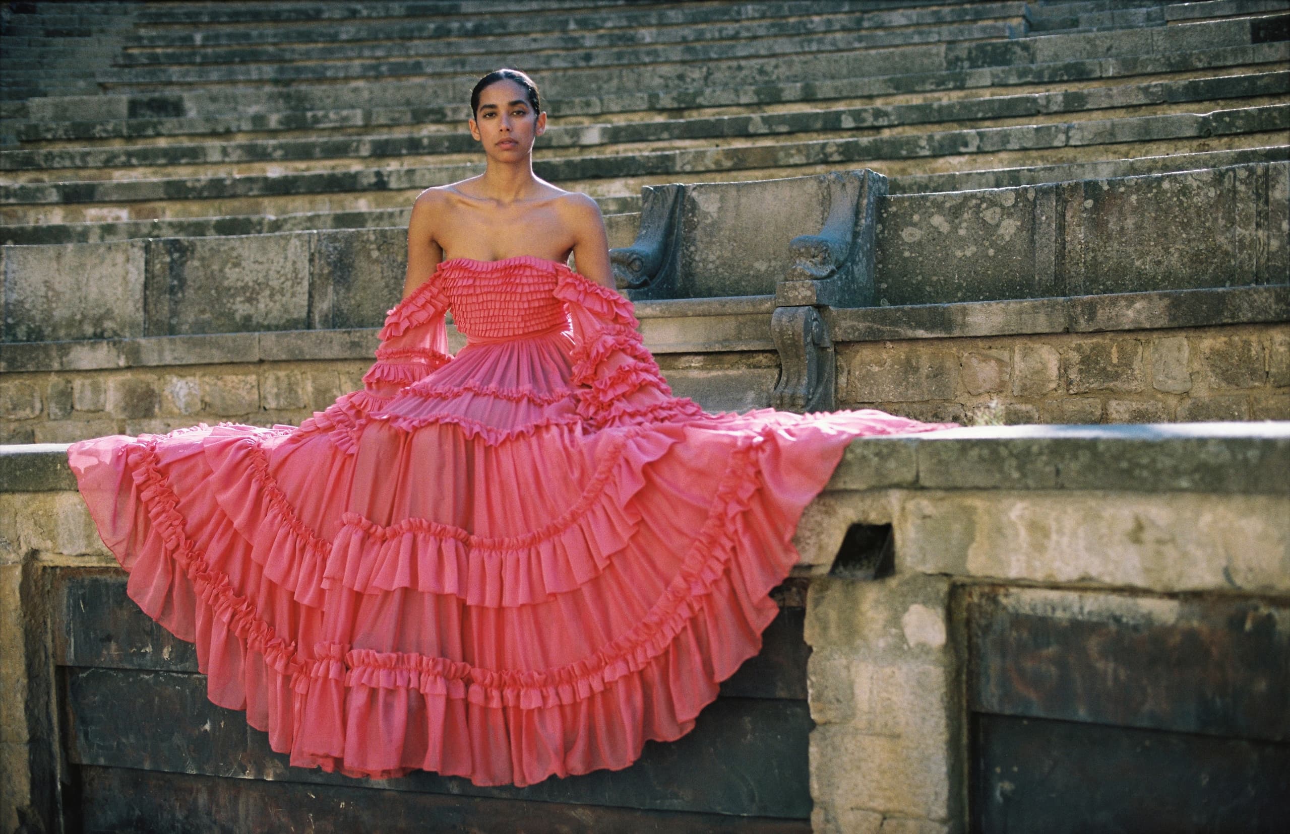 Woman in a pink ruffled gown sits on stone steps, serene and elegant.