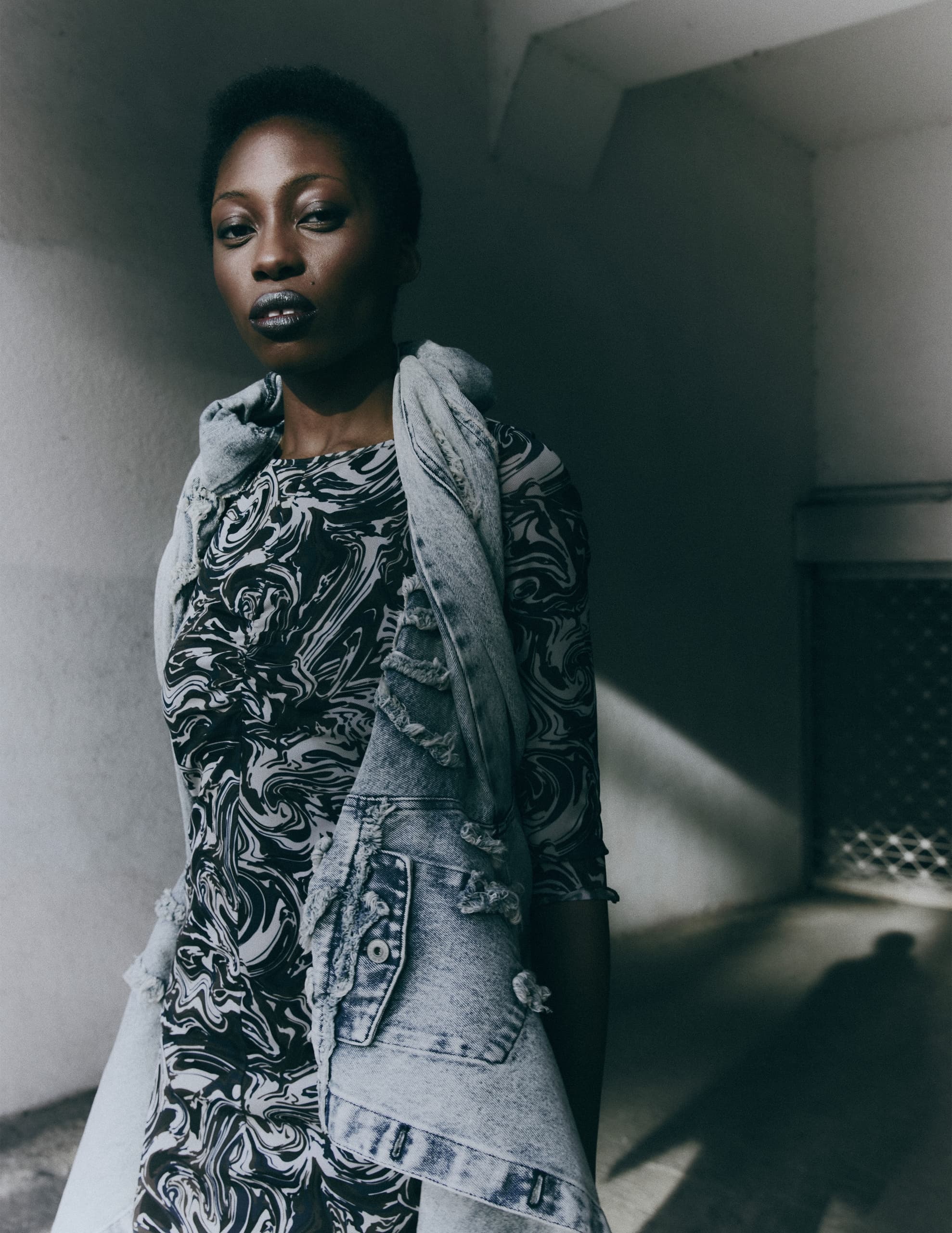 Fashionable person in patterned dress and denim vest, standing in moody lighting.