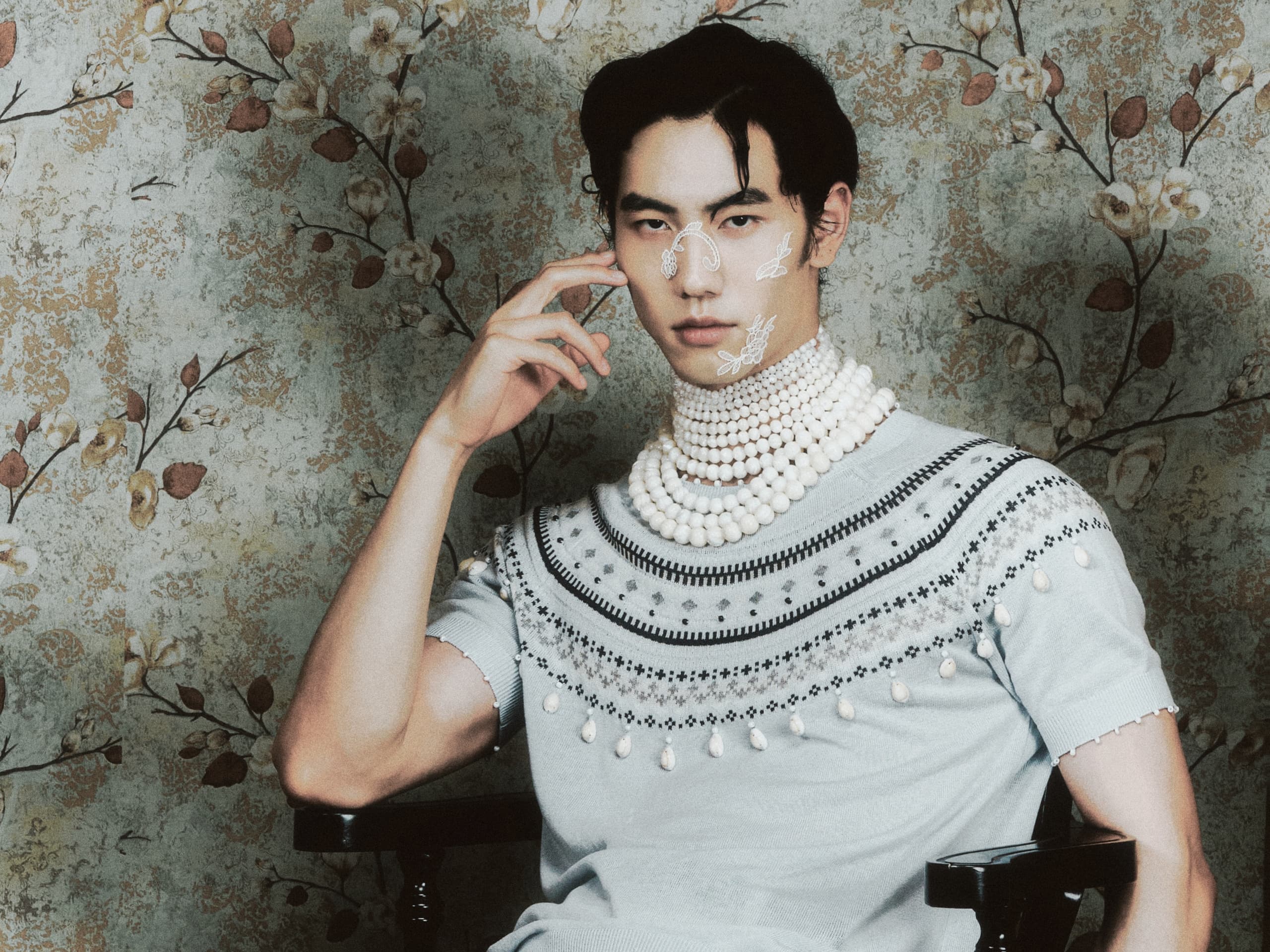 Fashion model with pearl accessories and embroidered top against floral wallpaper.