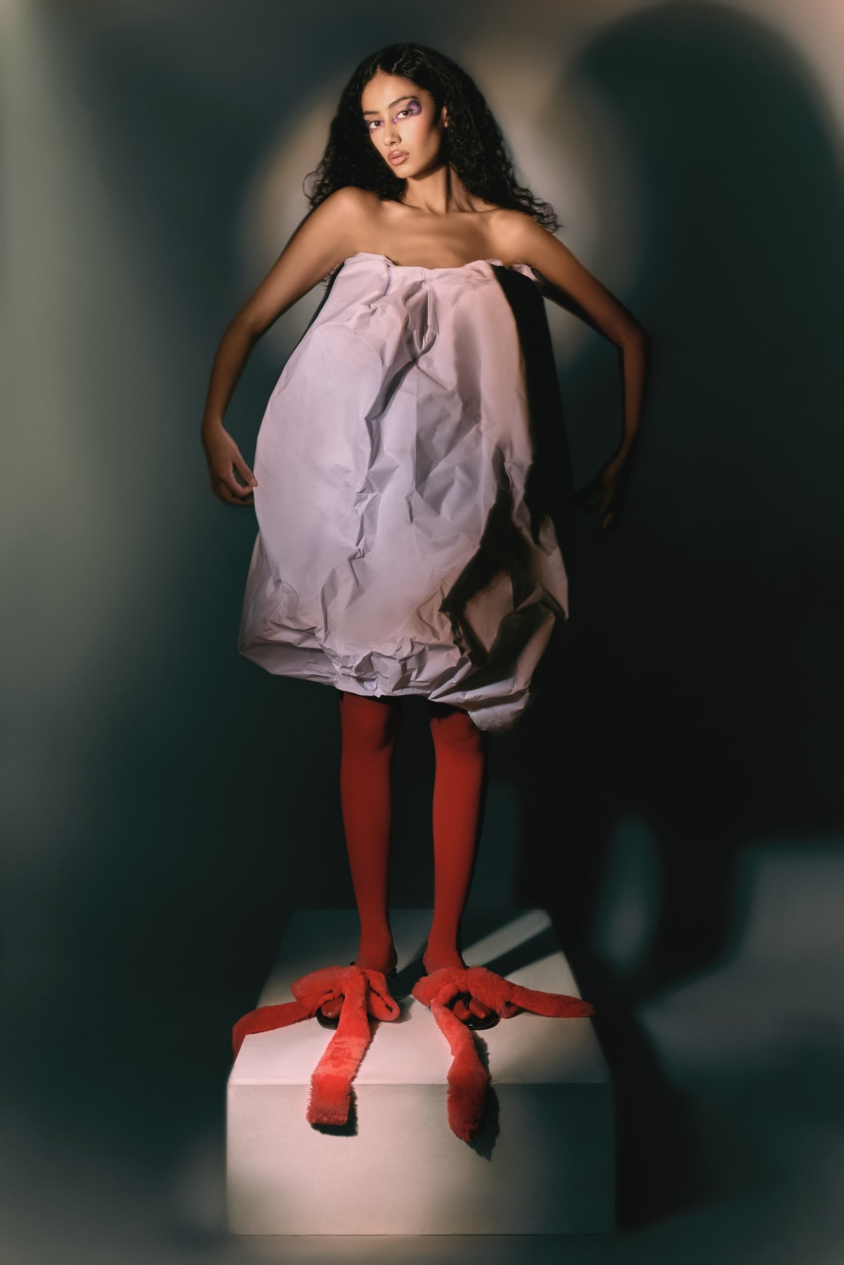 Fashion-forward model in creative white dress and bold red footwear, dramatic lighting.