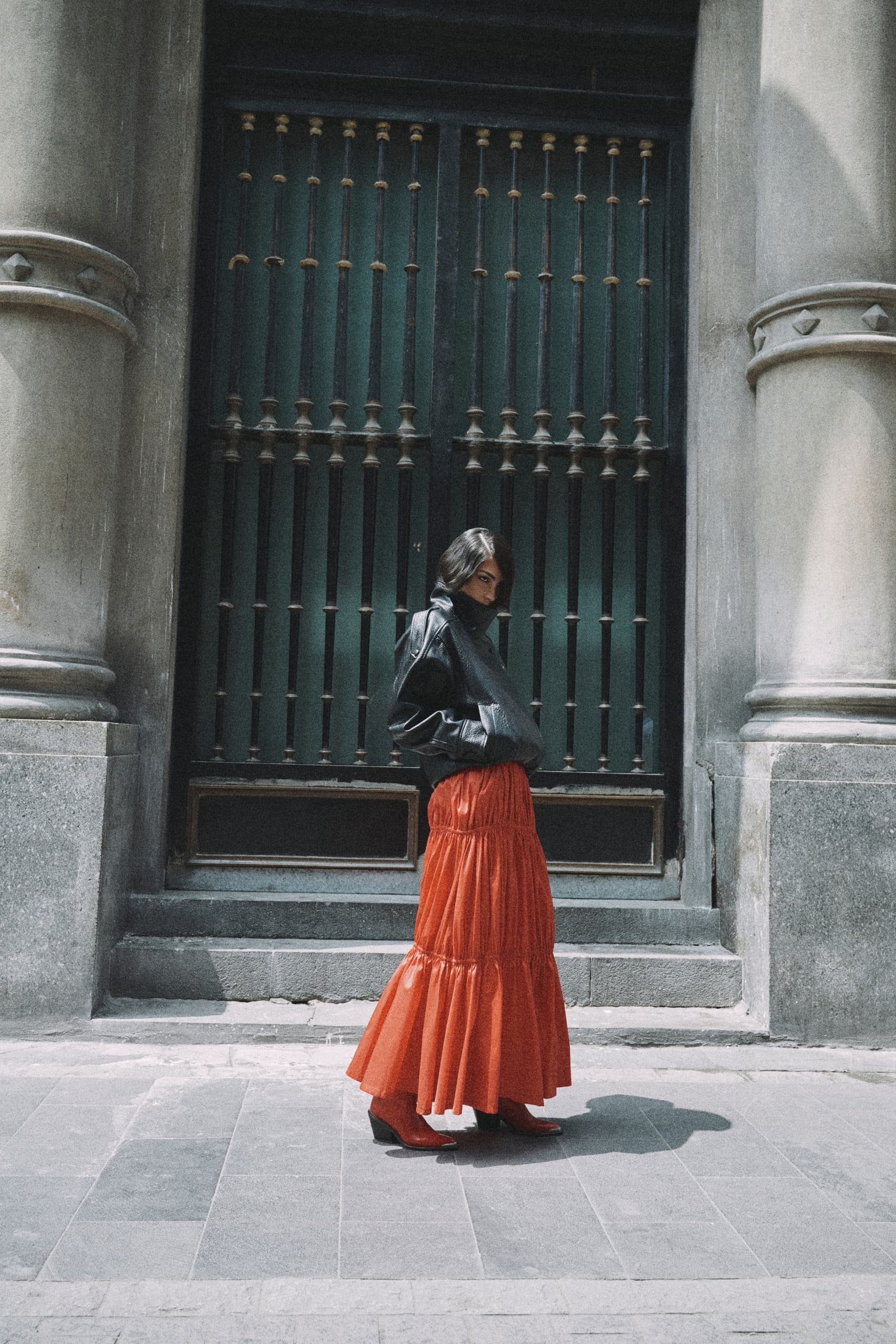 Woman in red skirt and black jacket standing before ornate gate.