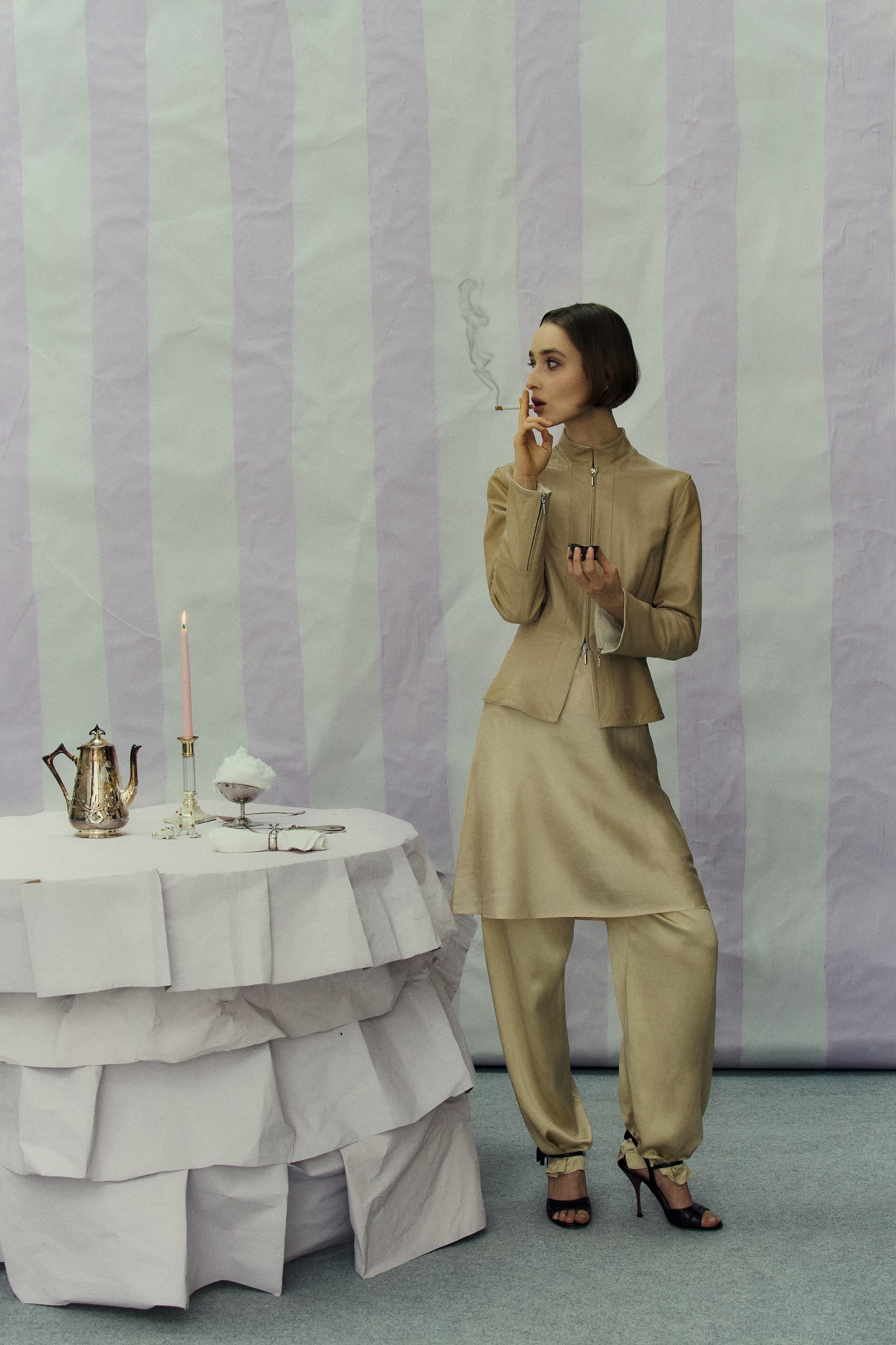 Elegant woman in beige outfit beside a table with candle and teapot, smoking.