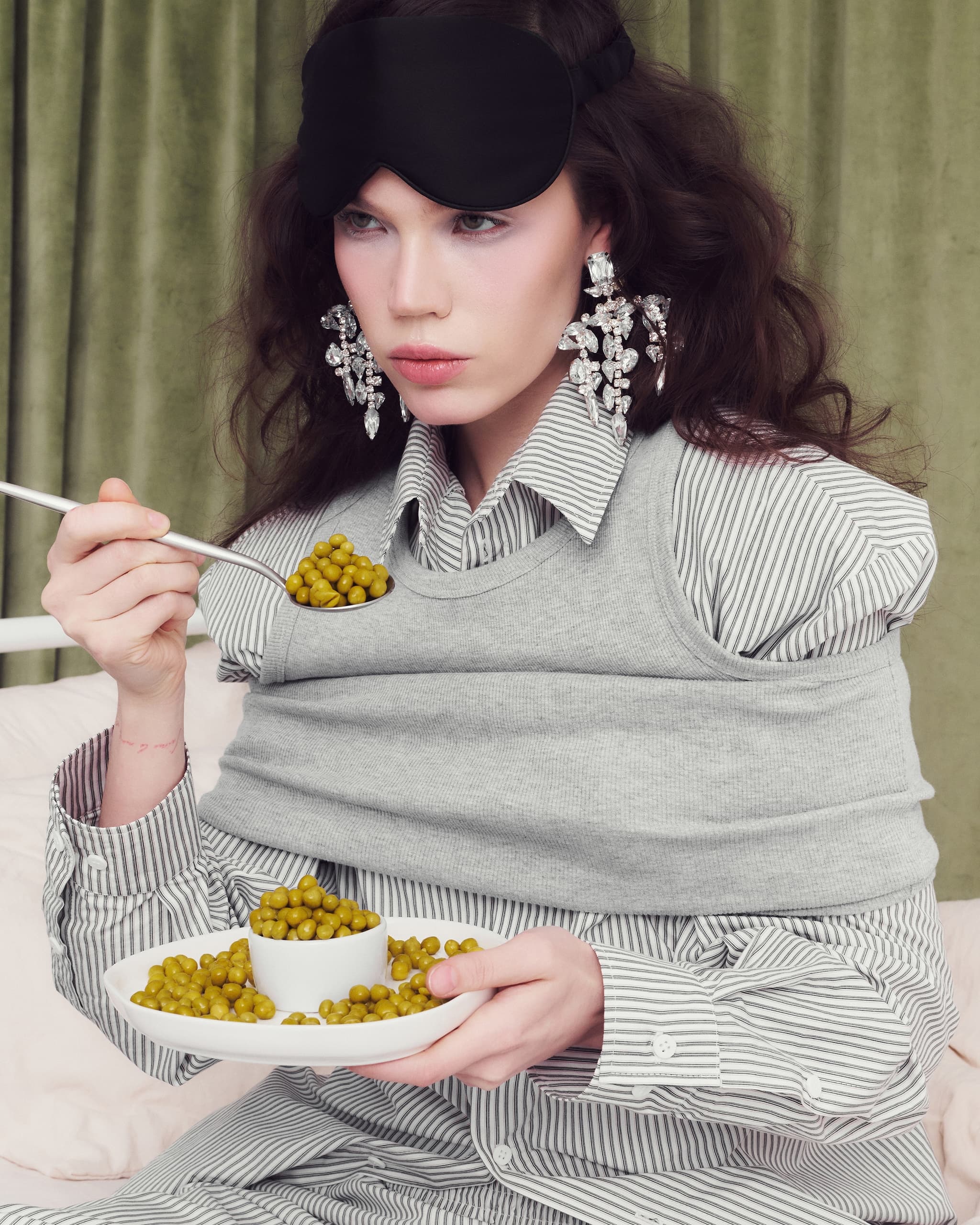 Fashionable woman eating peas; quirky style with eye mask and statement earrings.