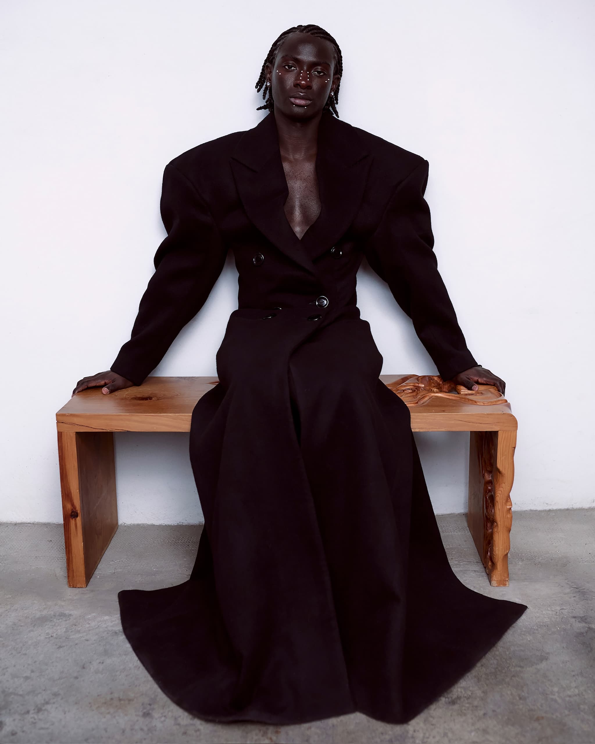 Stylish individual in a dramatic long black coat seated on a wooden bench.