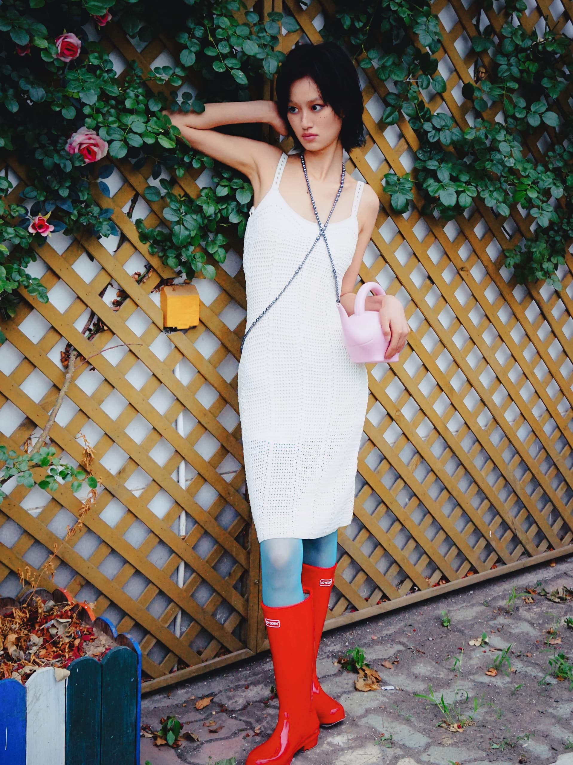 Woman in a white dress and red boots holding a pink watering can by a lattice fence.