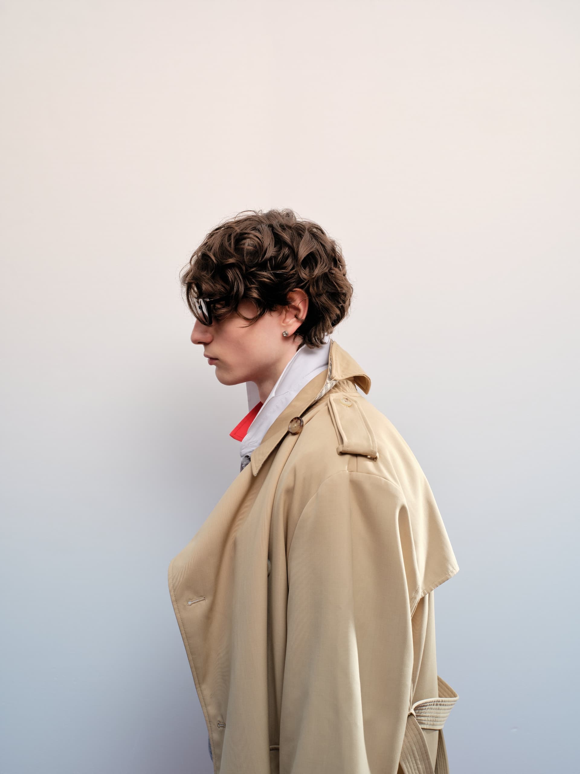 Person wearing trench coat and glasses in profile view against a plain background.