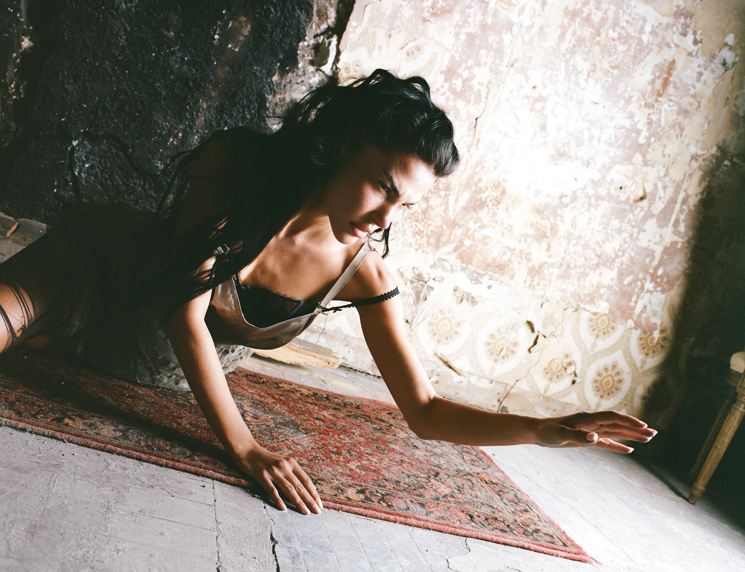 Woman in vintage dress stretching on patterned rug in rustic, textured room.