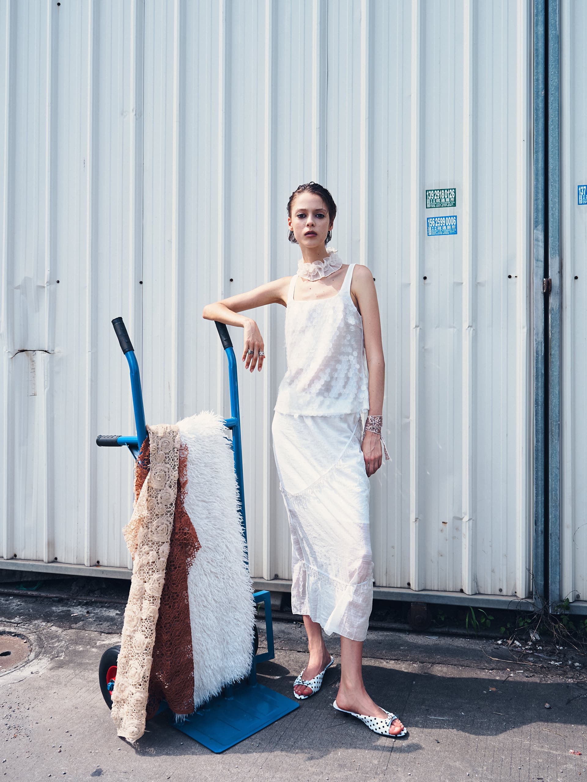 Woman in chic white outfit posing beside textiles and dolly cart outdoors.