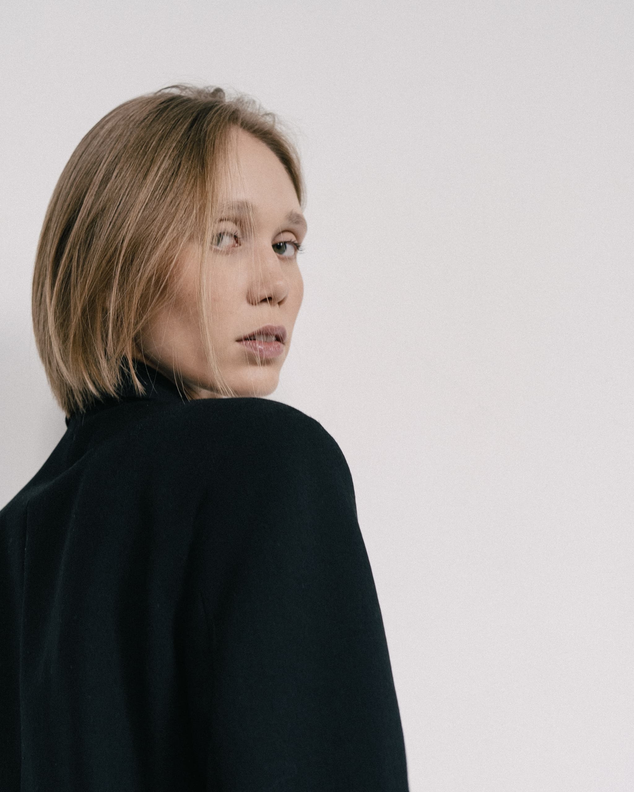 Person wearing black coat gazes back against white background.