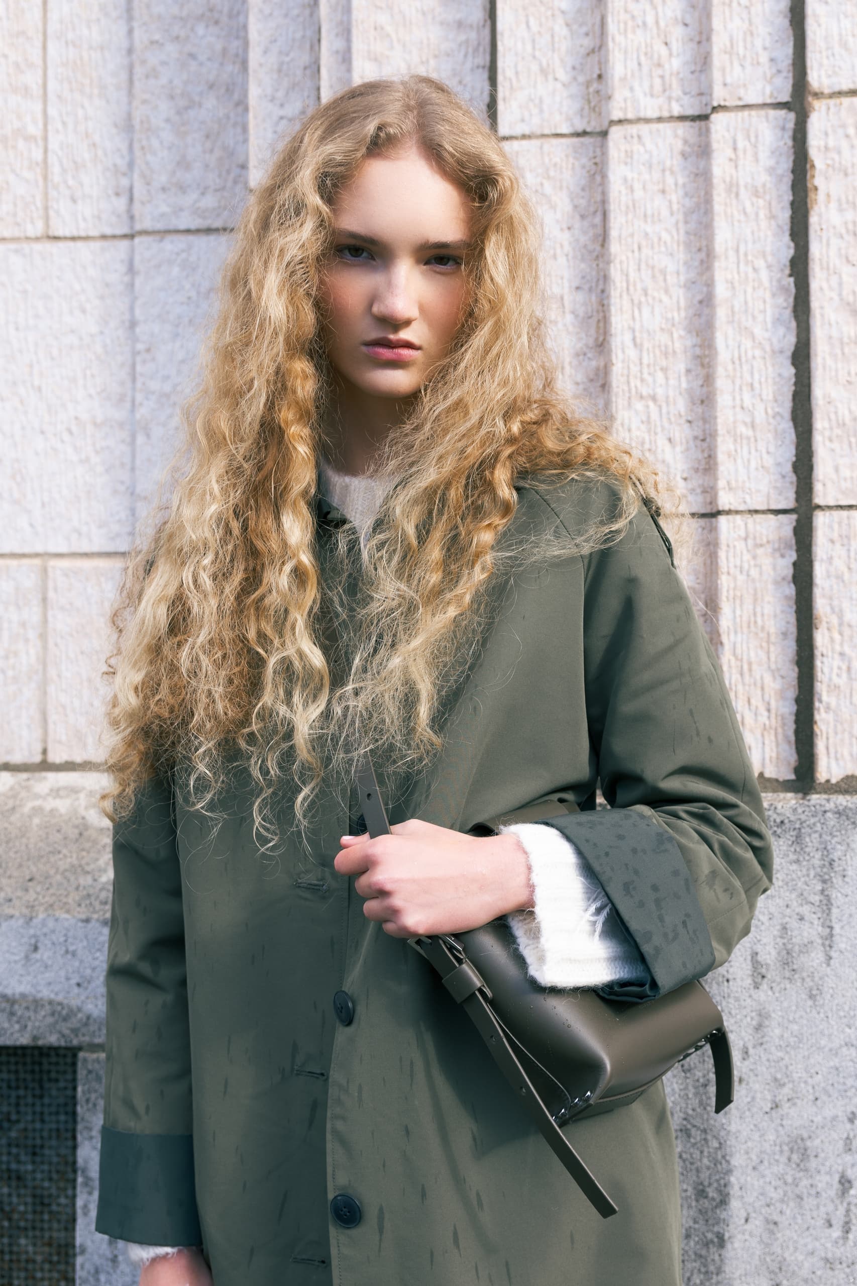 Woman in olive coat with long curly hair standing against a textured stone wall.