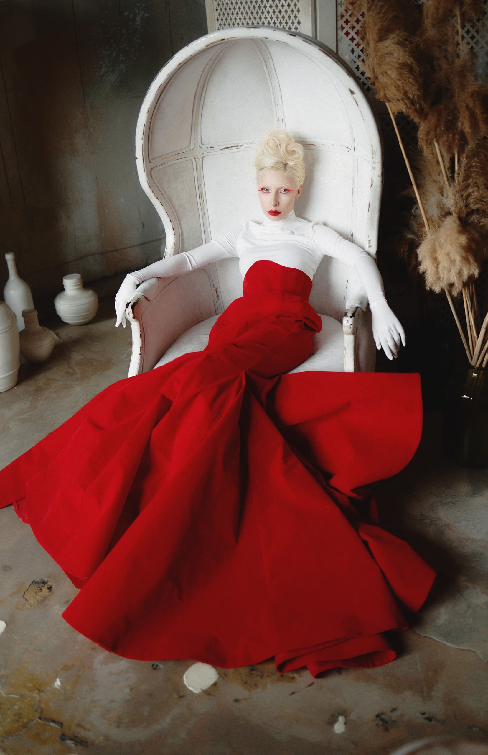 Elegant woman in red gown and white chair, striking high-fashion pose.