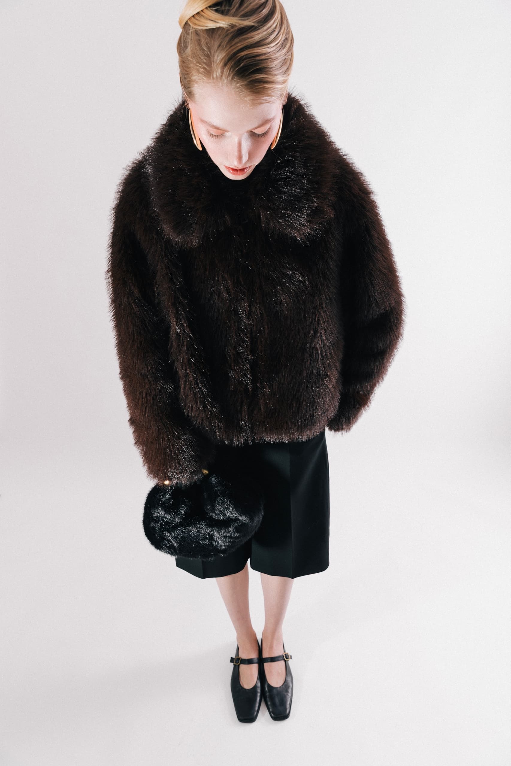 Woman in elegant fur coat with black purse and heels on white background.