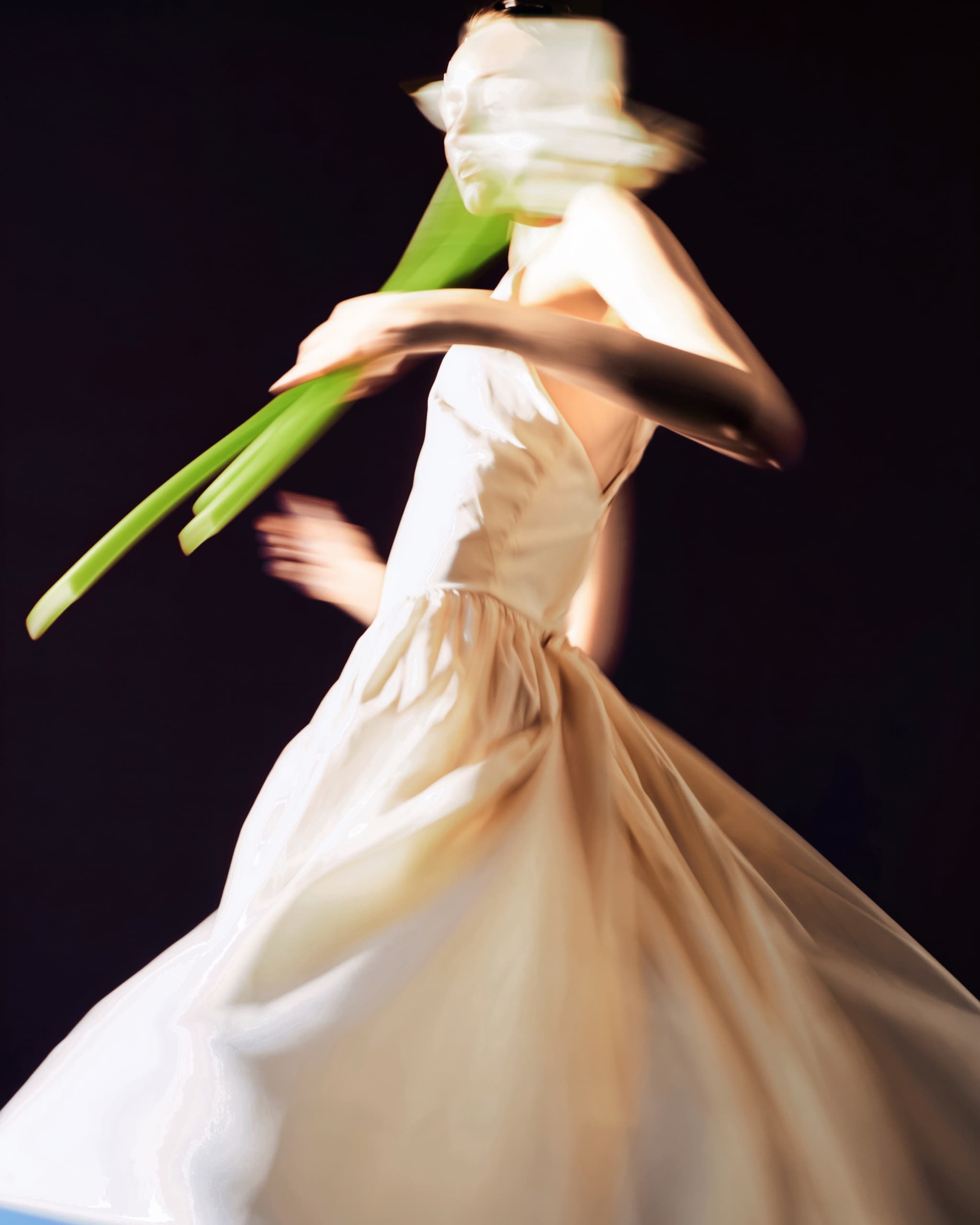Blurred motion of woman in flowing dress holding green stems, dark background.