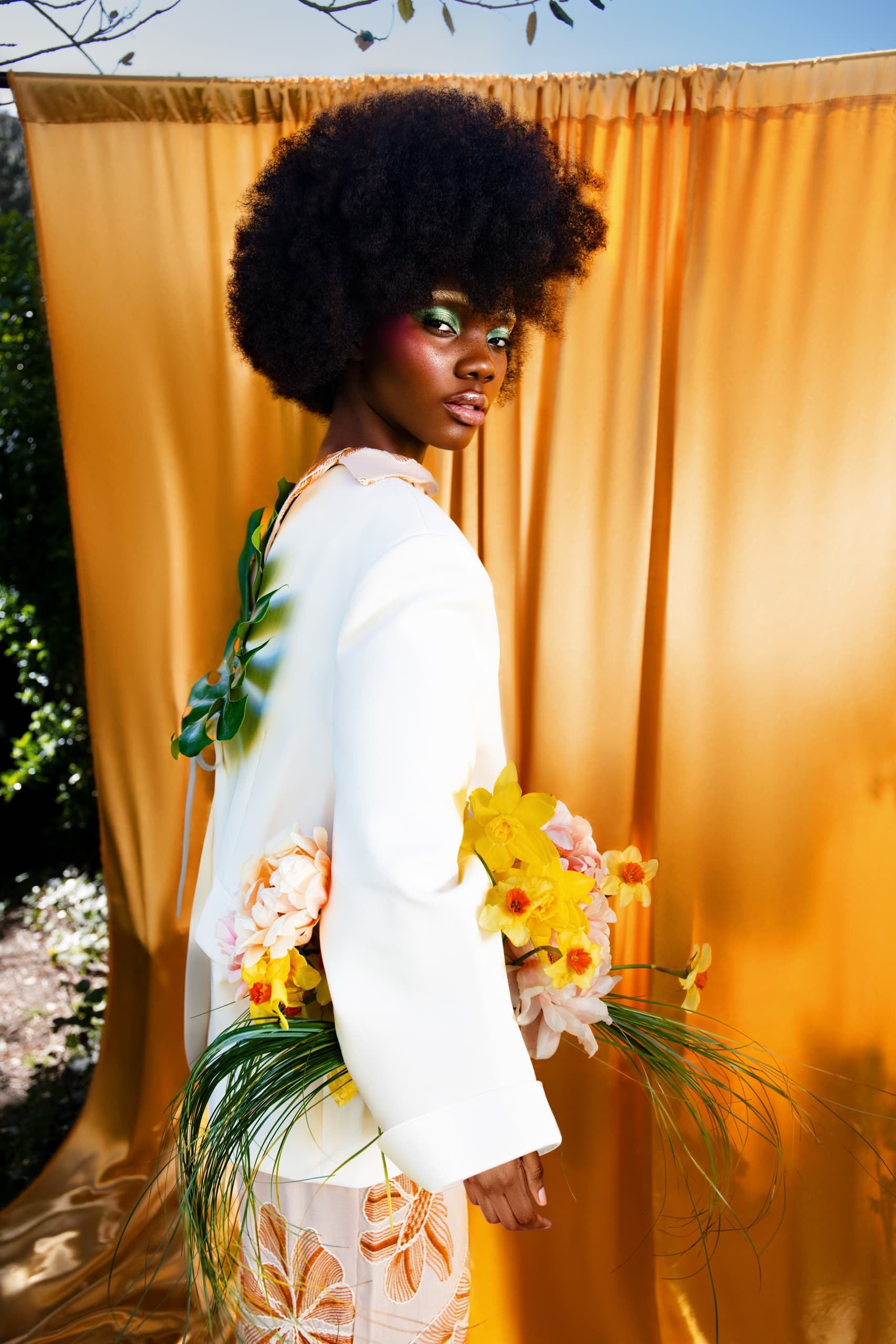 Fashionable woman in white coat with afro hairstyle holding vibrant flowers outdoors.