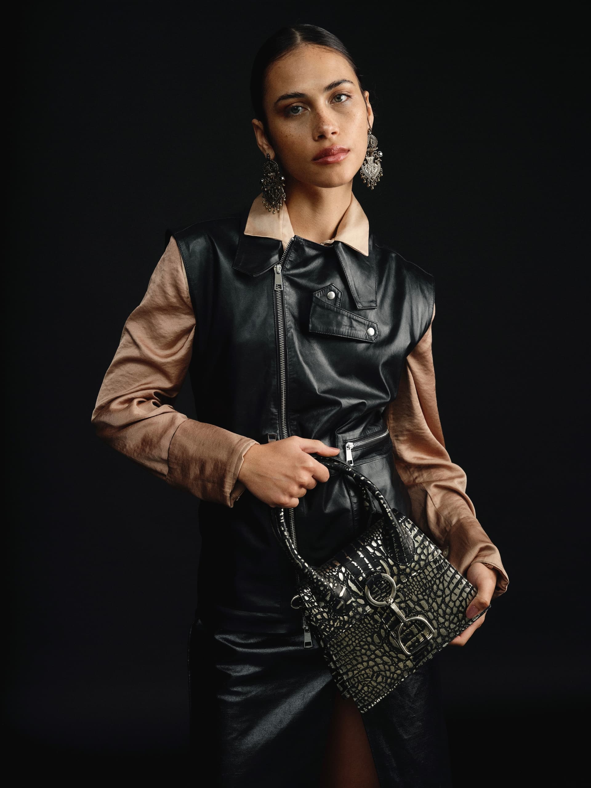 Woman in stylish outfit holding a crocodile-patterned handbag, dark backdrop.