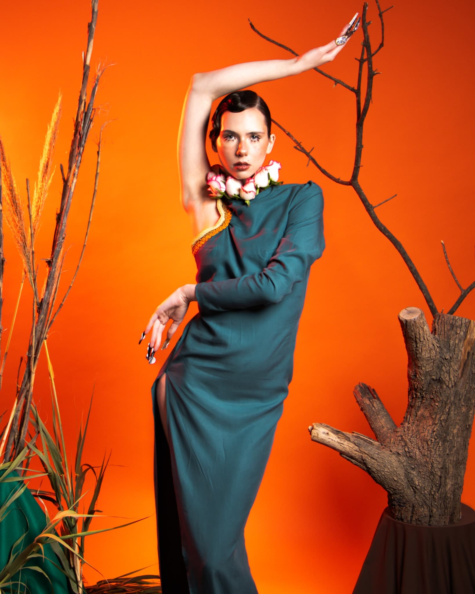 Model in teal dress with floral neckpiece posing, orange backdrop, artistic scene.