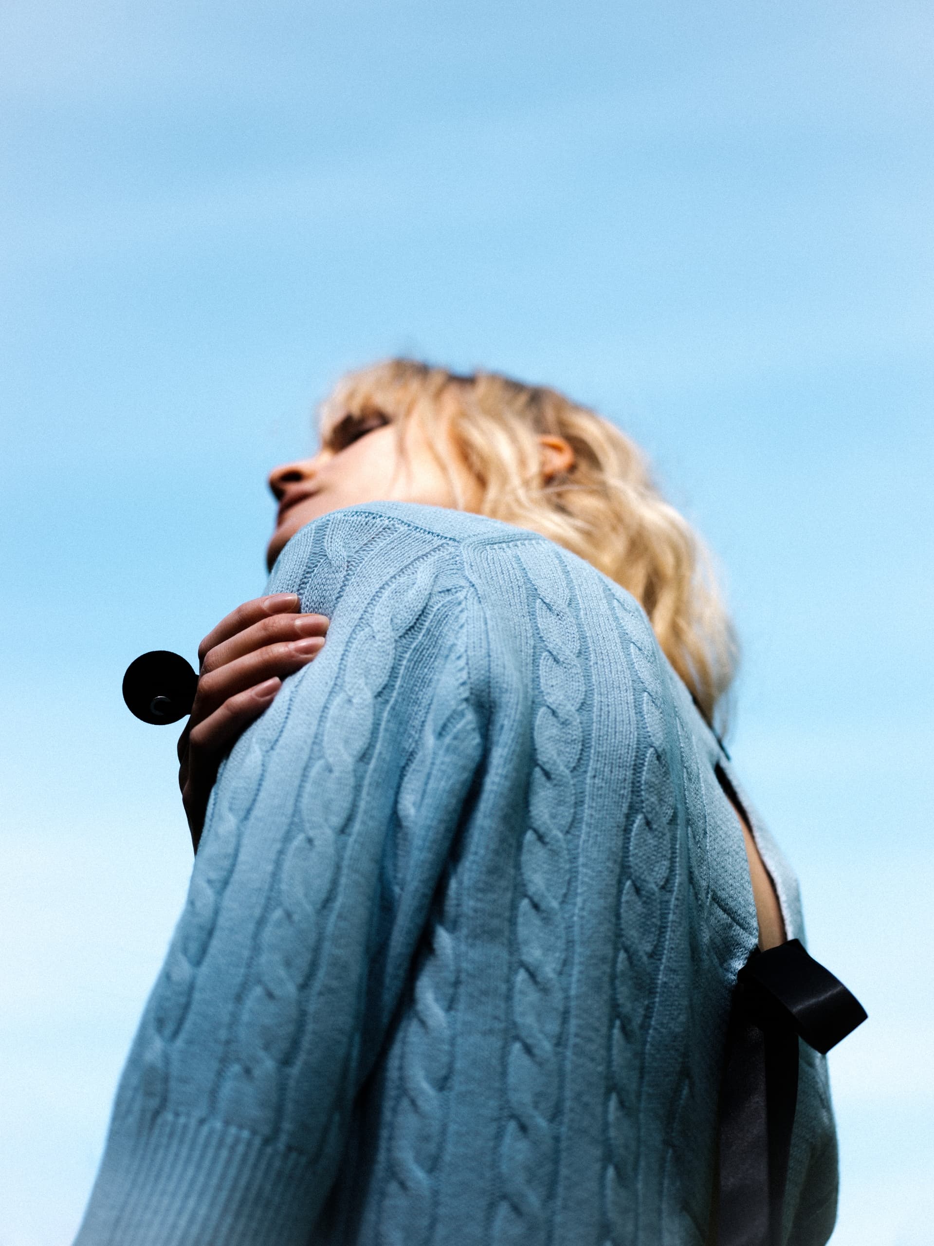 Person in blue sweater gazing at sky, holding object, under clear blue sky.
