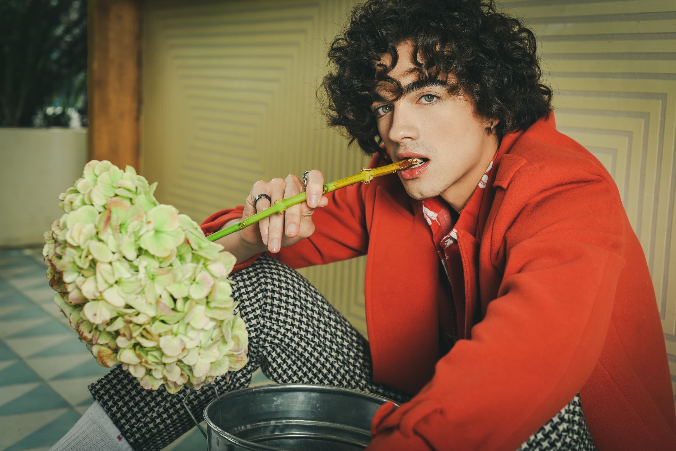 Curly-haired person in red coat holding hydrangea in a stylish pose.