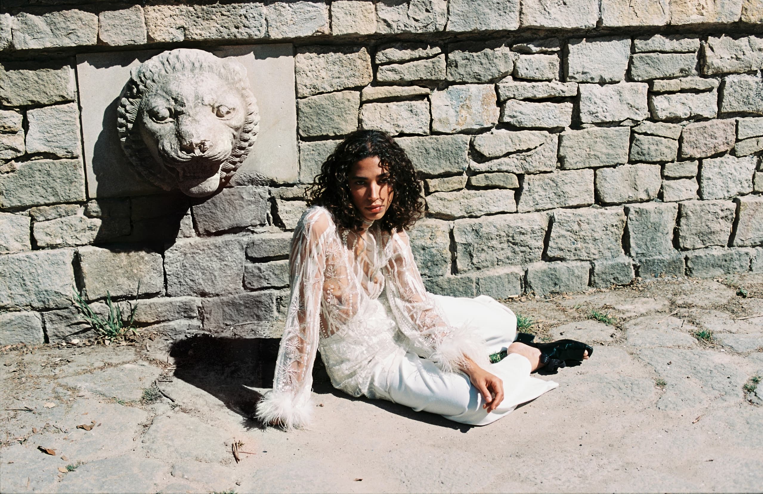 Person in stylish white outfit sitting by stone wall with sculpted lion head.