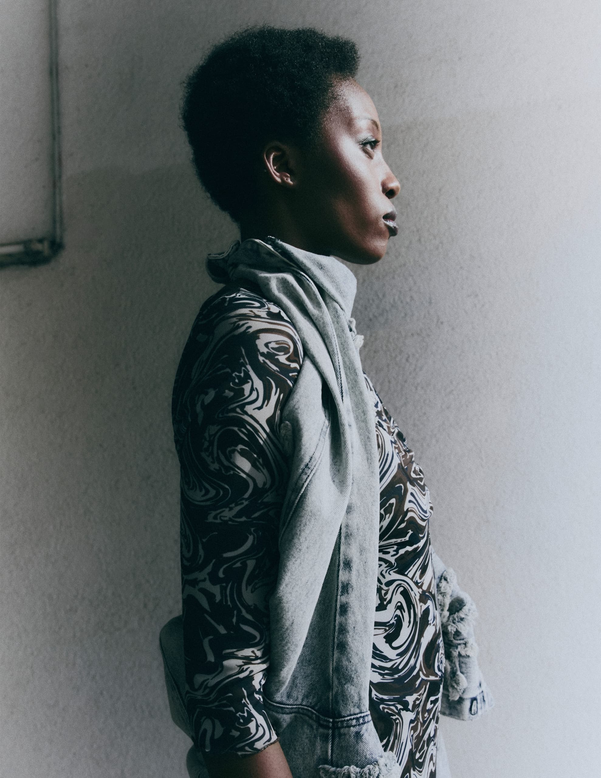Stylish woman in profile, wearing patterned shirt and denim jacket against white wall.