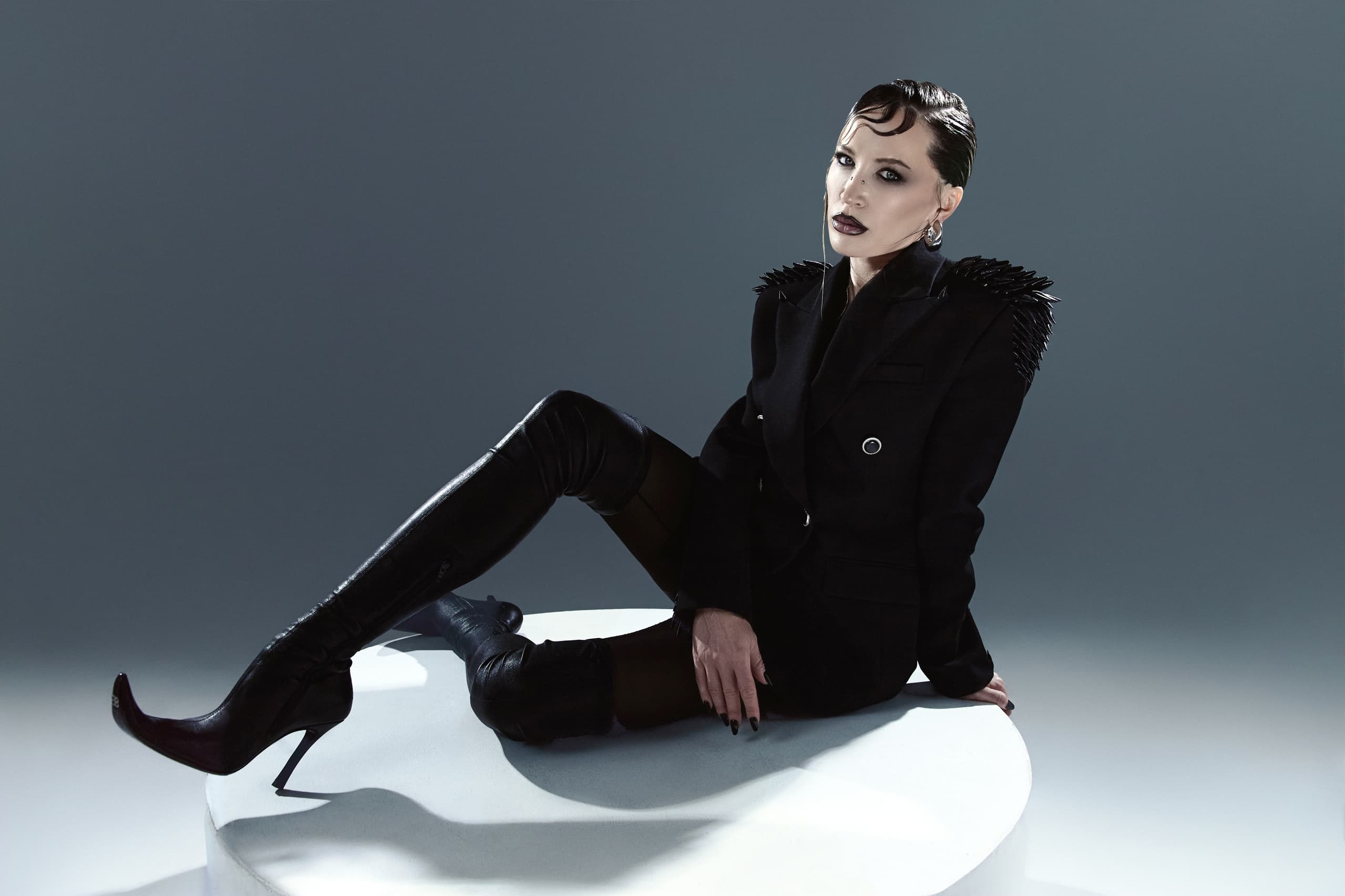 Fashionable model in edgy black outfit and high boots sitting on a white platform.