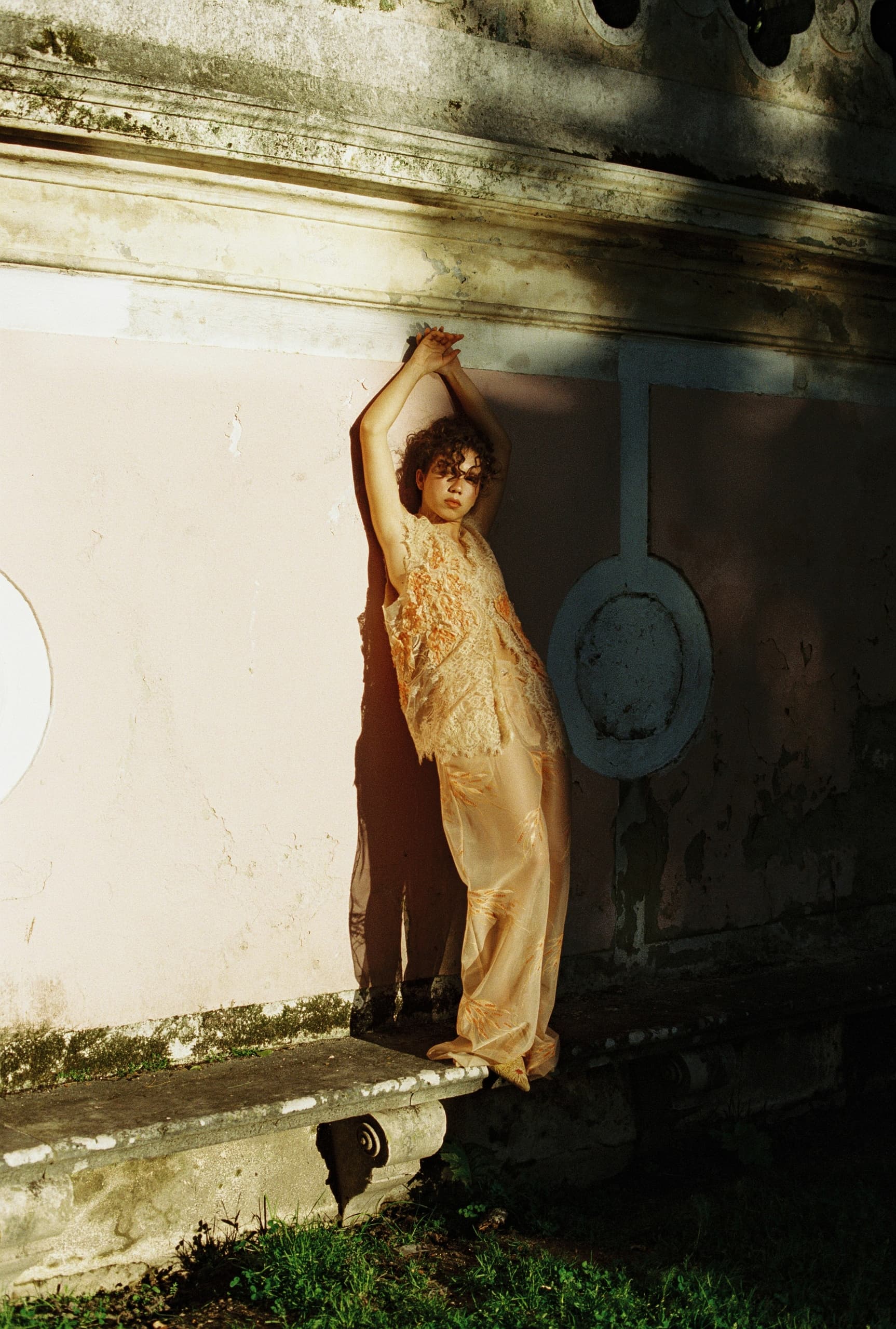 Person in sheer outfit leaning against an ornate wall in natural sunlight.