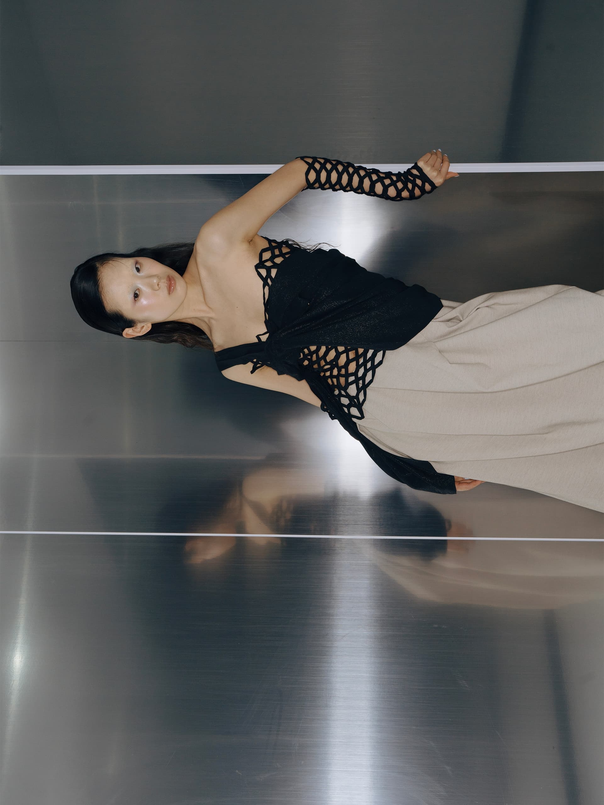 Fashionable woman poses in chic netted black top and beige skirt against a metal wall.