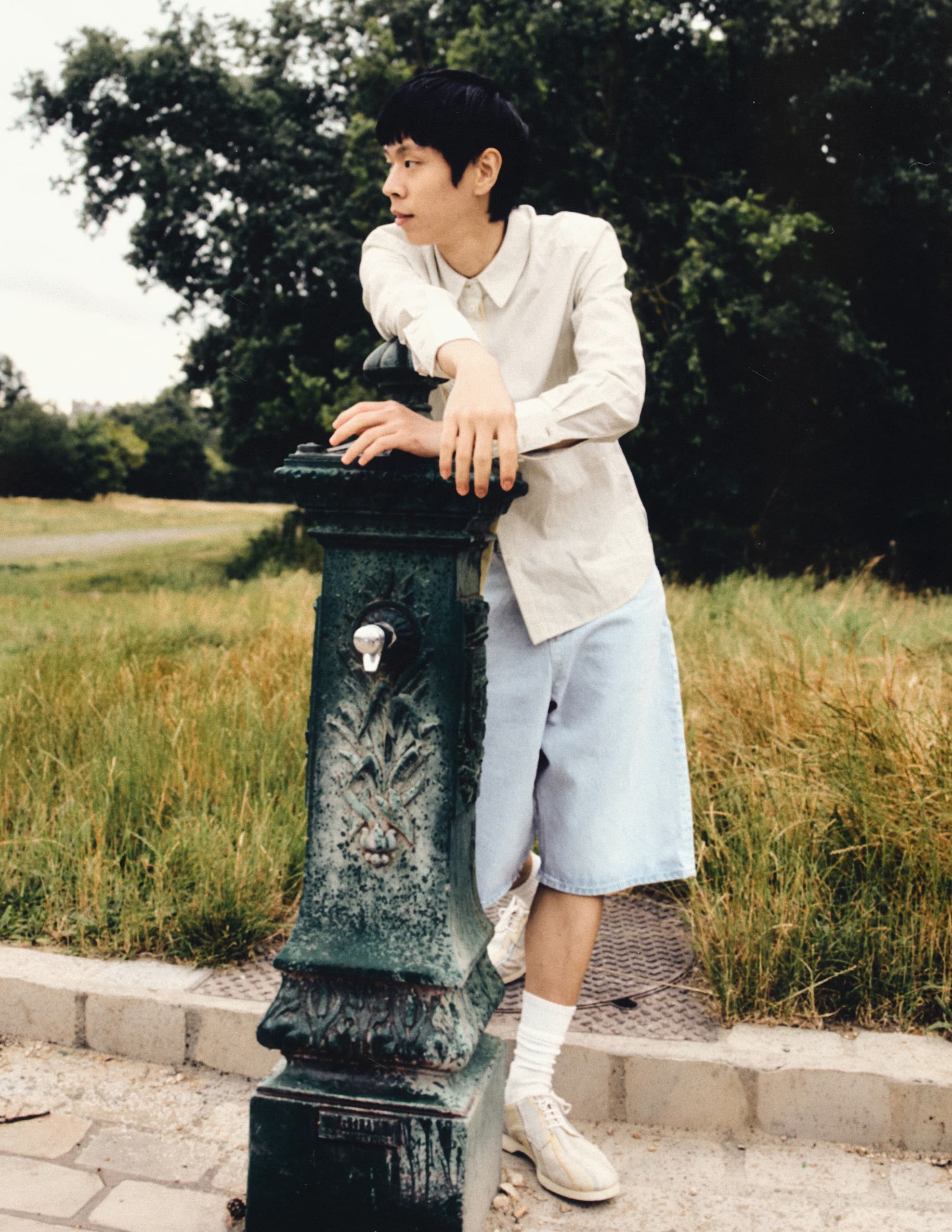 Person leaning on a vintage water fountain in a park setting.