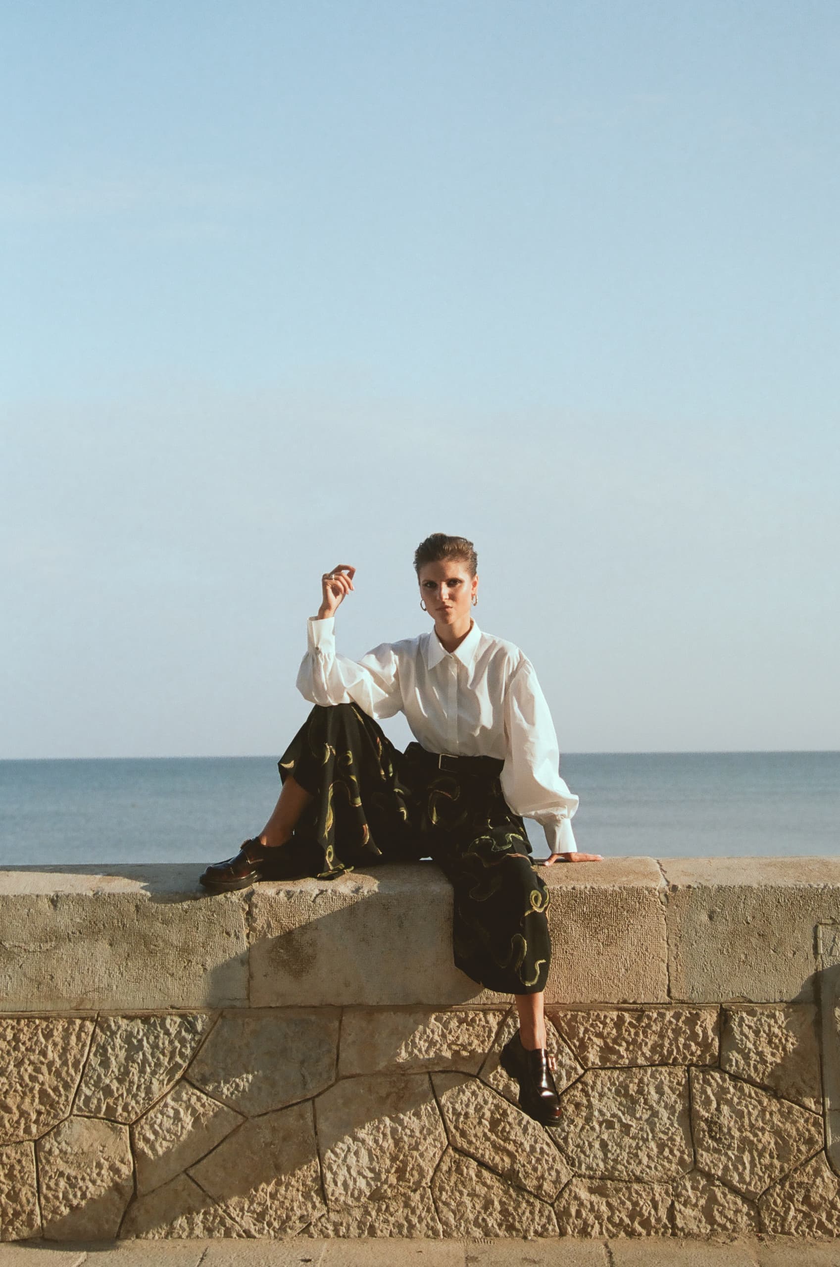 Person in stylish outfit sitting on seafront wall, calm ocean background.